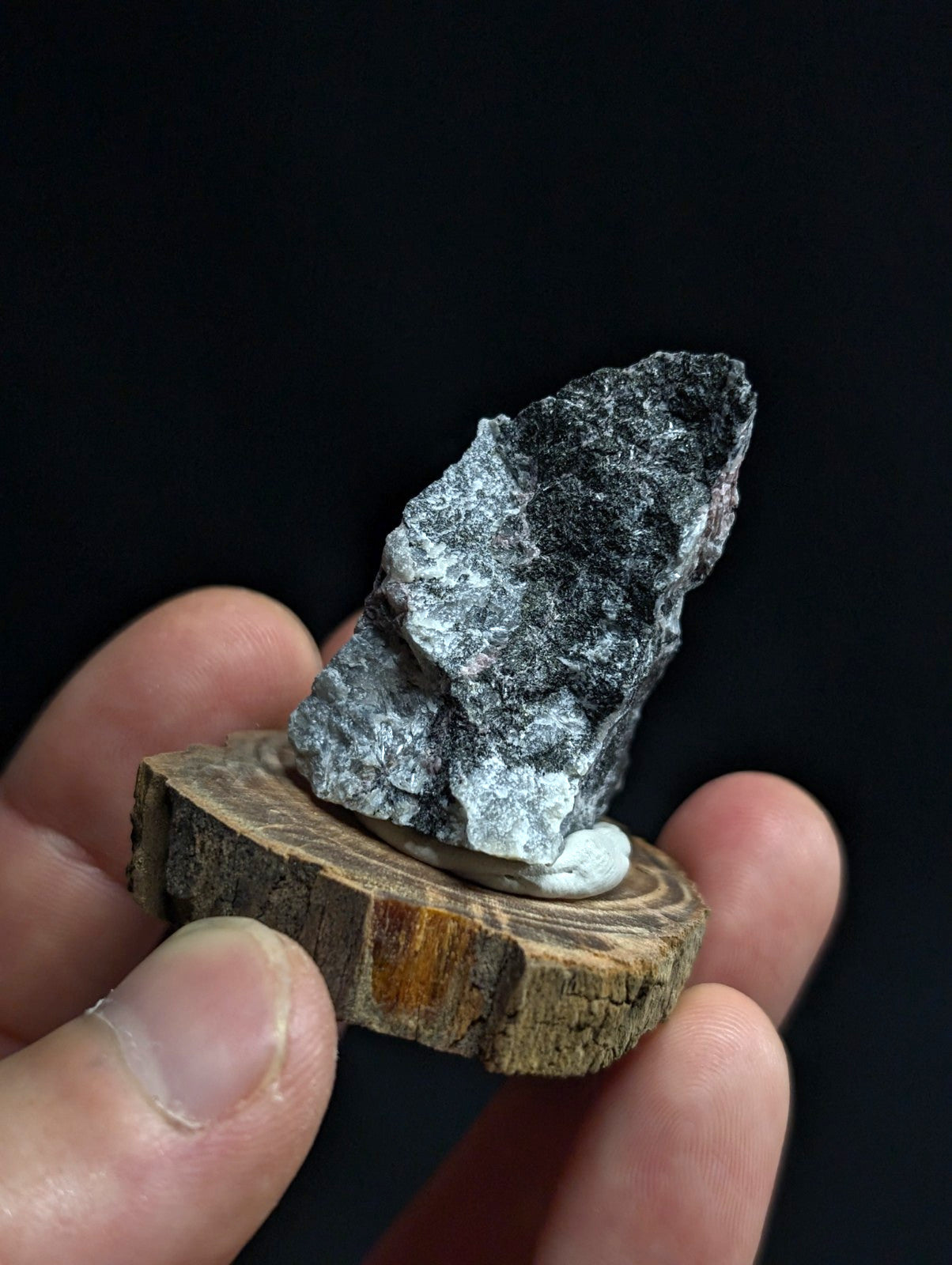 A rugged, naturally occurring mineral specimen with a mottled gray and black surface, displayed on a rustic wooden slice.