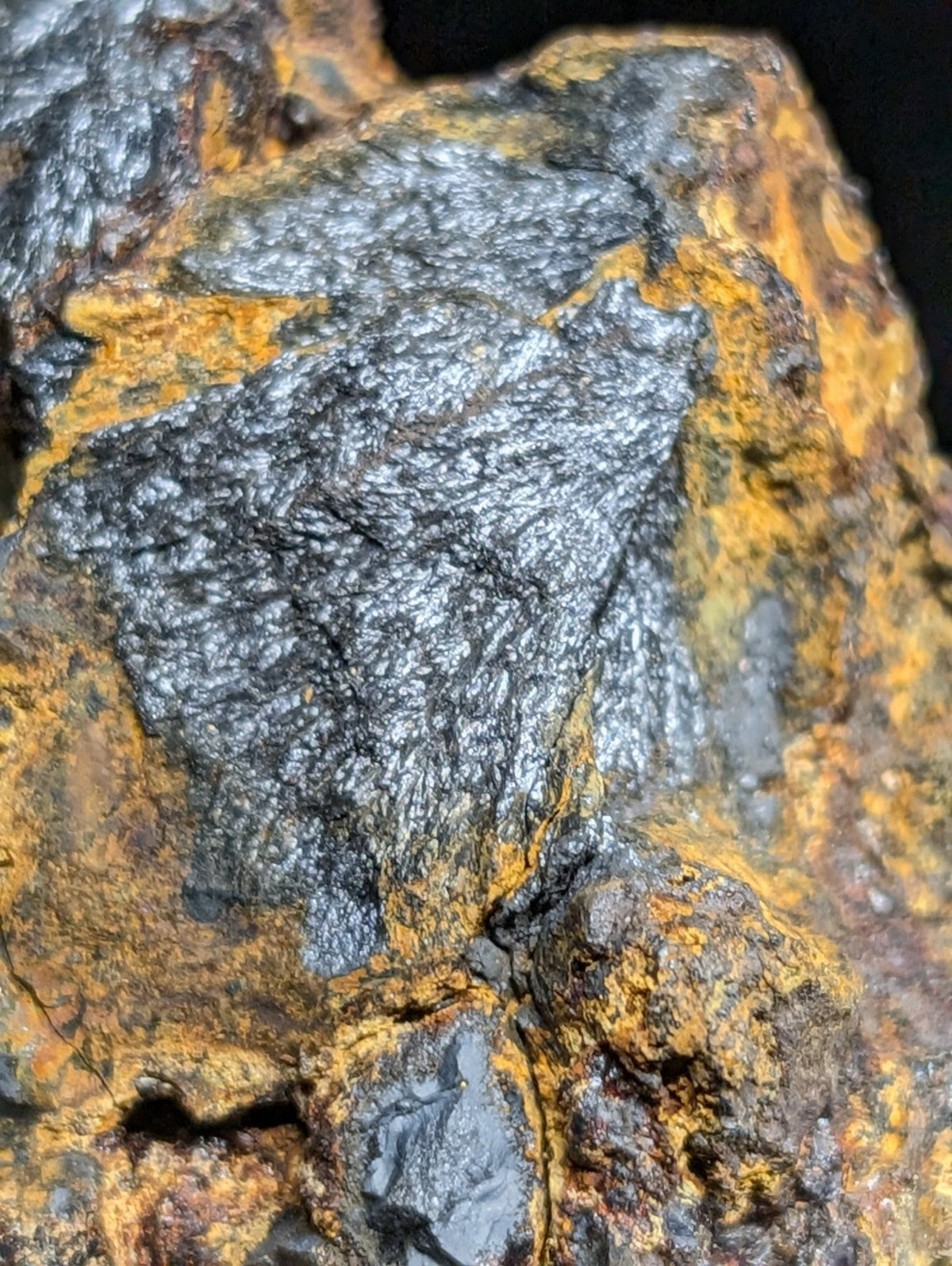 A rugged, dark metallic mineral specimen with a textured surface and patches of vibrant orange-yellow oxidation.