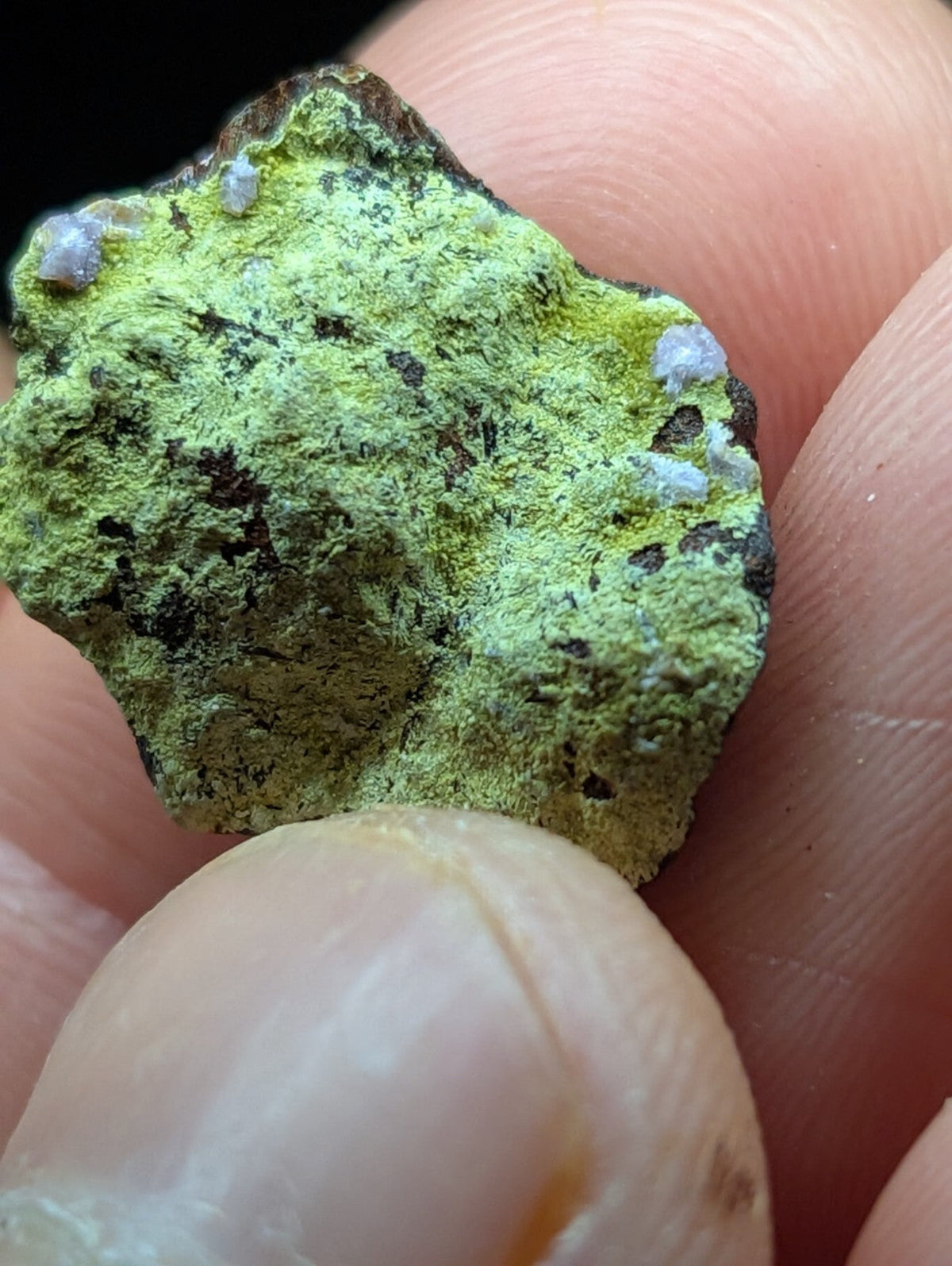 A rough, irregularly shaped mineral specimen with a vibrant green surface speckled with dark patches and small white crystalline formations.