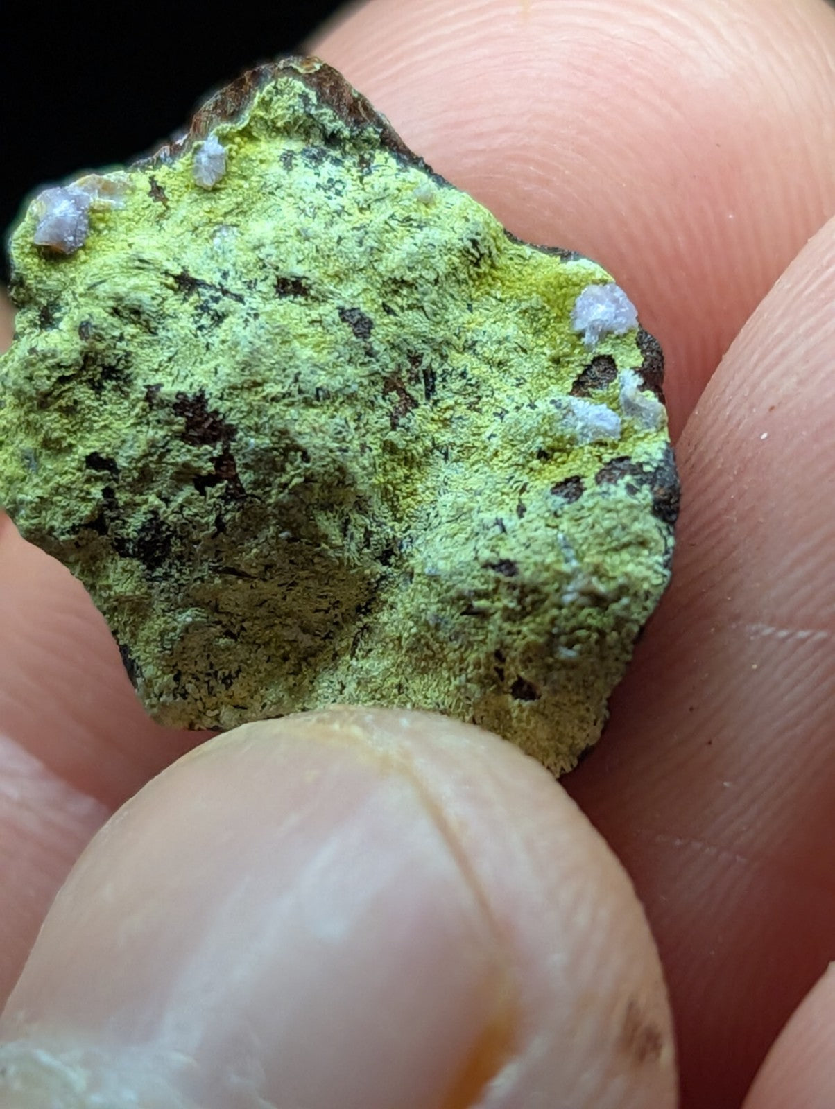 A rough, irregularly shaped mineral specimen with a vibrant green surface speckled with dark patches and small white crystalline formations.