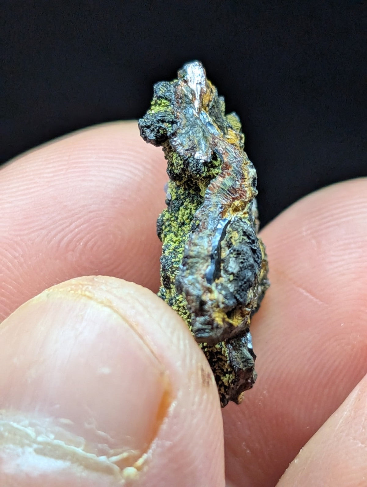 A rugged, irregularly shaped mineral specimen with a dark metallic base accented by patches of green and yellowish hues.