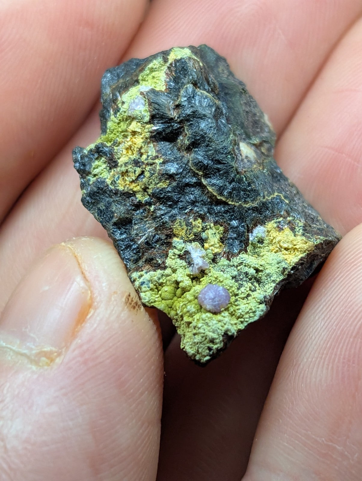 A rough, irregularly shaped mineral specimen with a dark base and patches of vibrant green patina, featuring a small purple gemstone embedded in its surface.