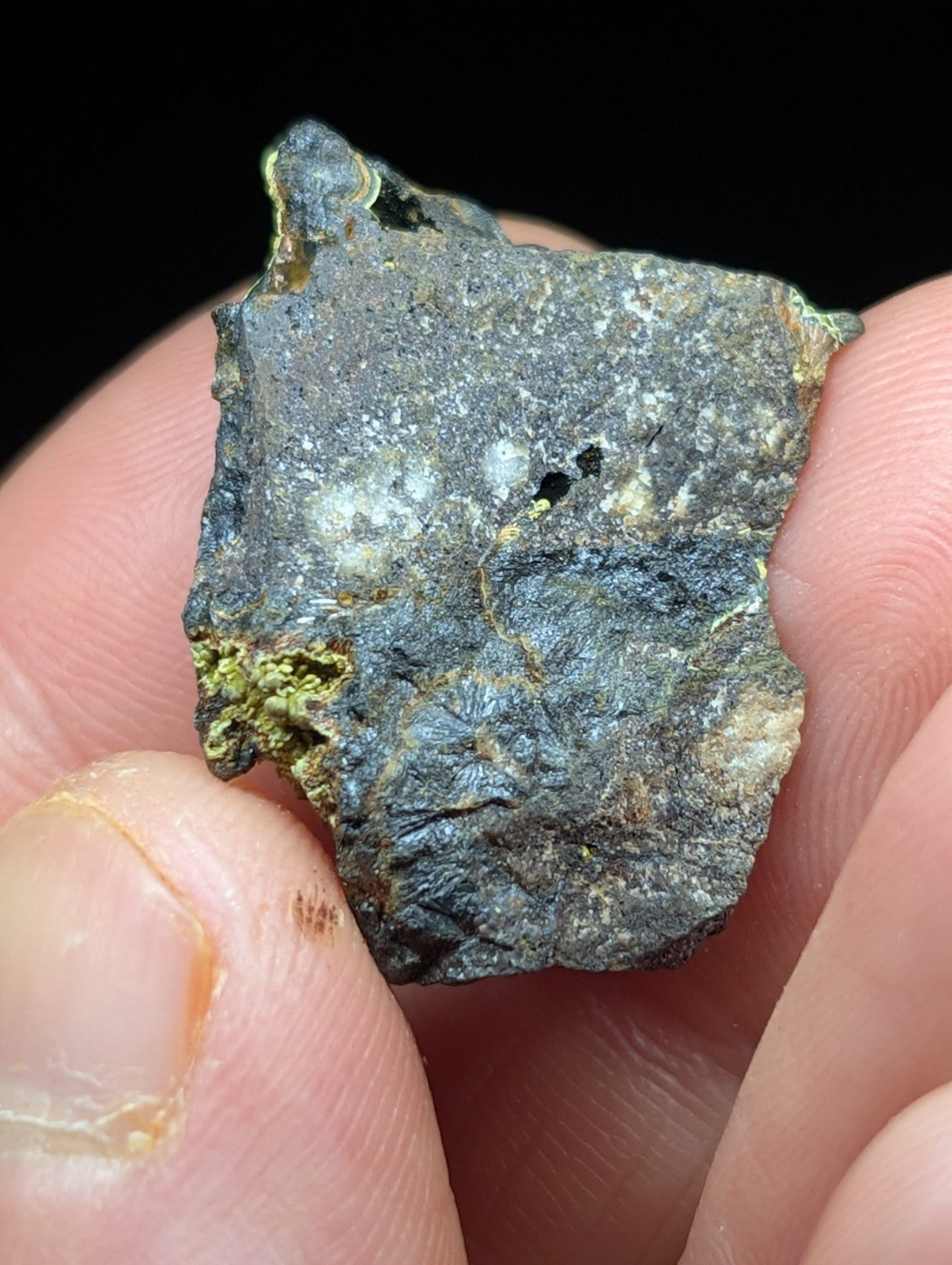 A rugged, irregularly shaped meteorite fragment with a dark gray surface marked by patches of greenish-yellow patina and subtle metallic sheen.