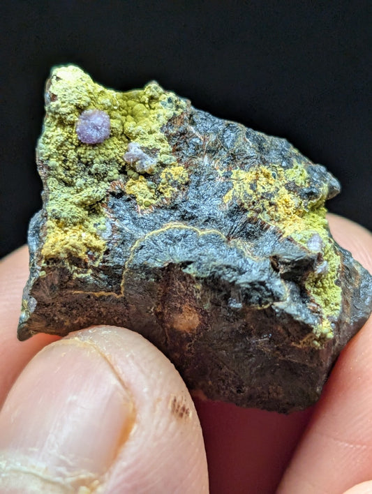 A dark, irregularly shaped mineral specimen with patches of vibrant green and yellow patina, featuring a small purple gemstone embedded in its surface.