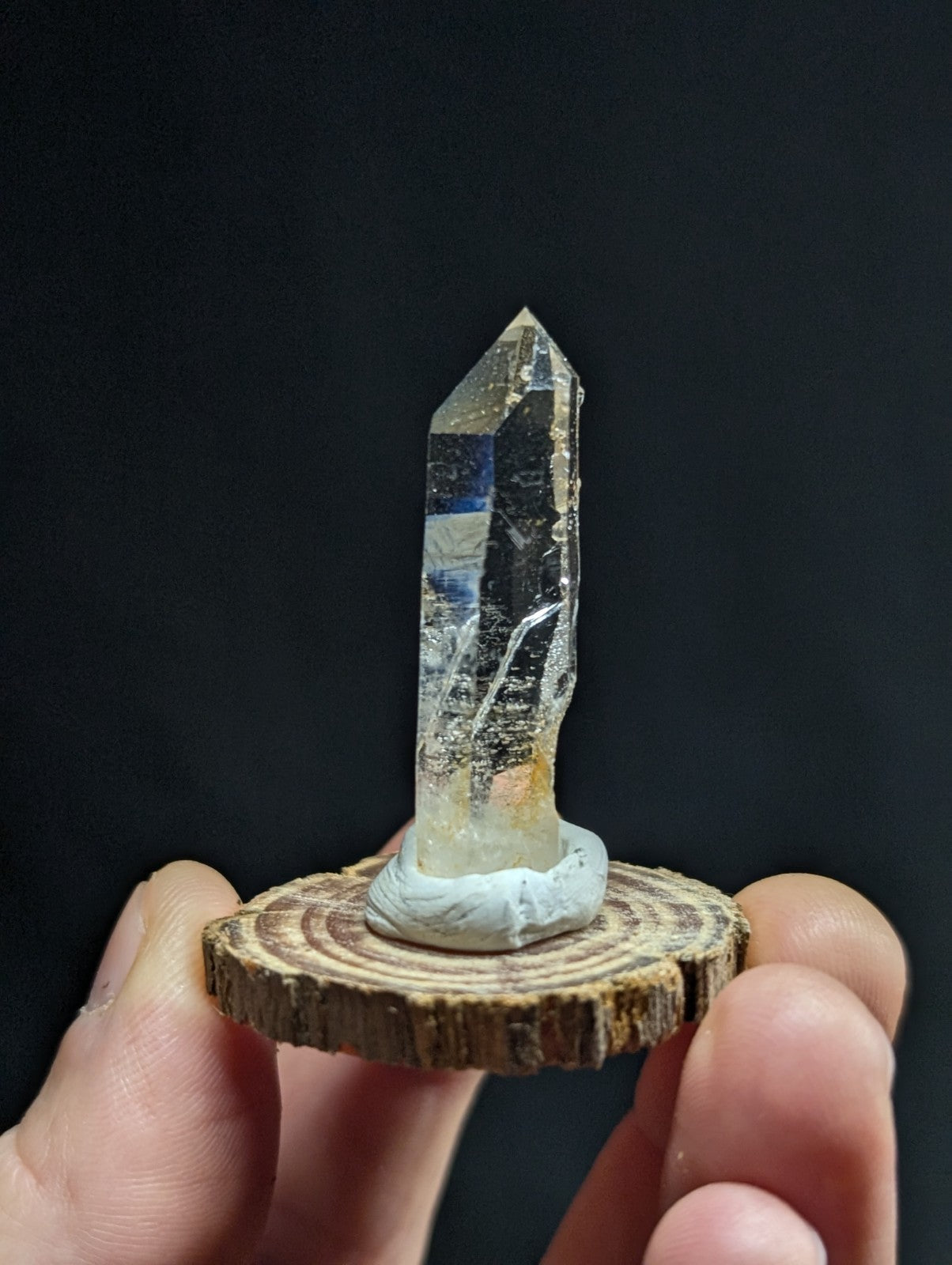 A clear quartz crystal with a pointed tip and natural inclusions, mounted on a circular wooden slice base.