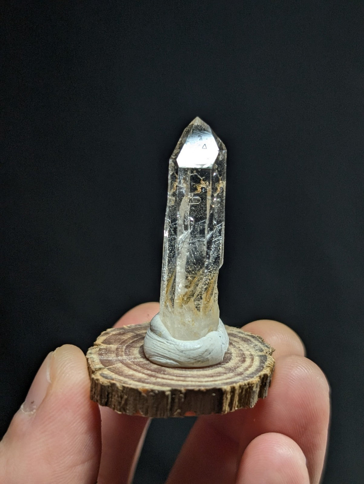 A clear quartz crystal with a pointed tip and natural inclusions, mounted on a rustic wooden slice base.