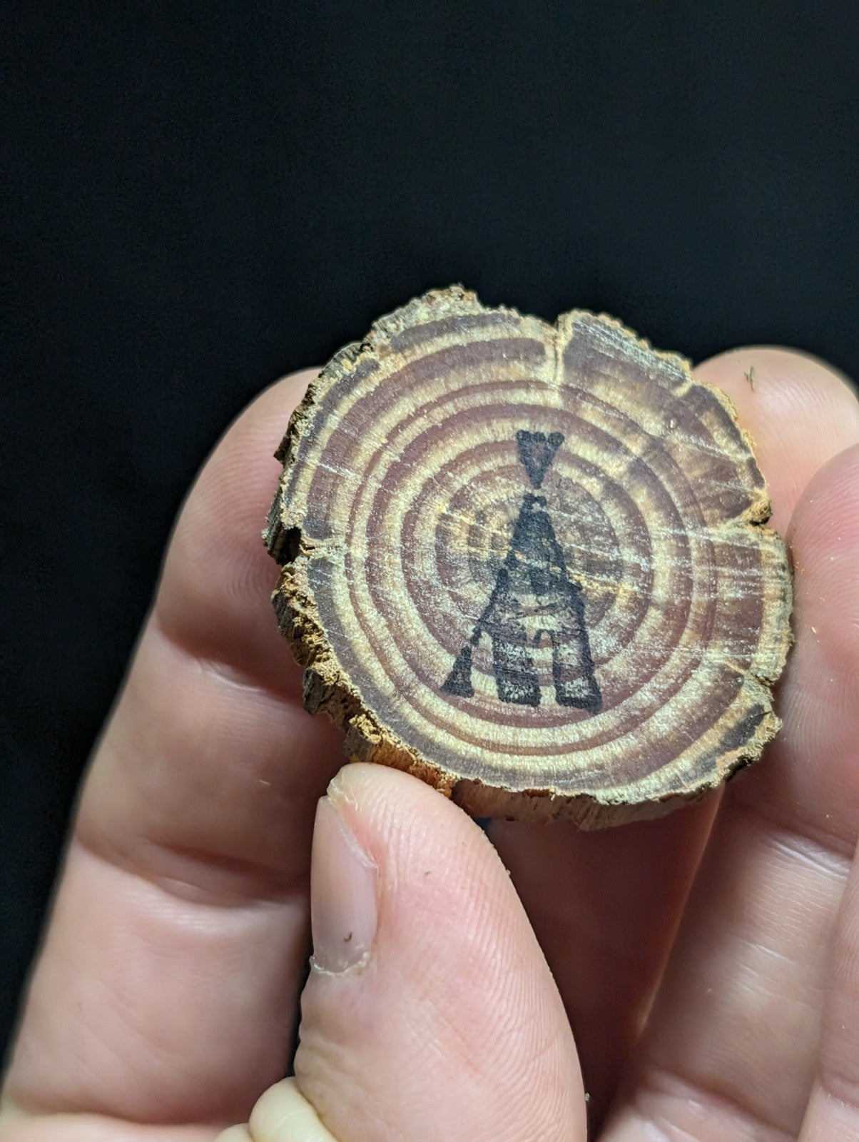 A circular wooden slice with natural brown rings and a black inked teepee design at its center.