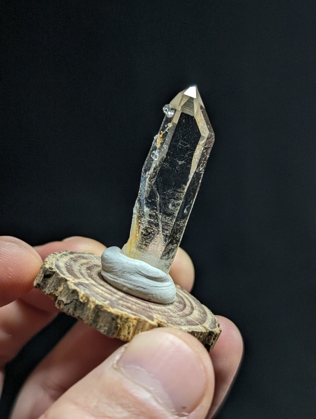 A clear, faceted quartz crystal with a pointed tip and subtle internal inclusions is held upright on a rustic wooden slice base.