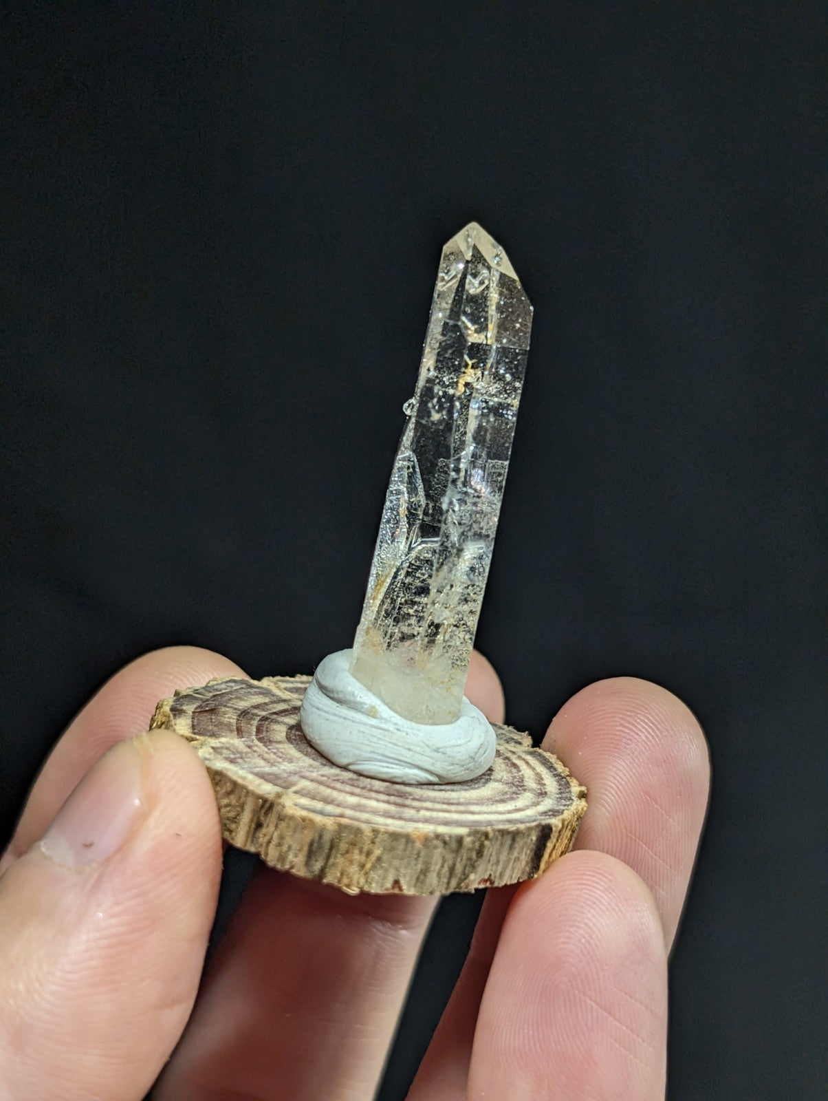 A clear quartz crystal with a pointed tip and natural inclusions, mounted on a circular wooden slice base.