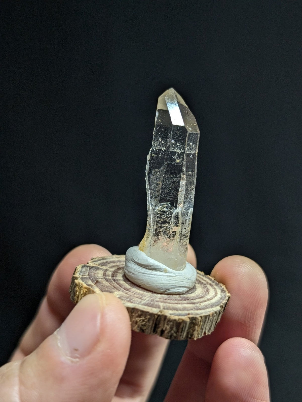 A clear quartz crystal with a pointed tip and visible internal striations, mounted on a circular wooden slice base.