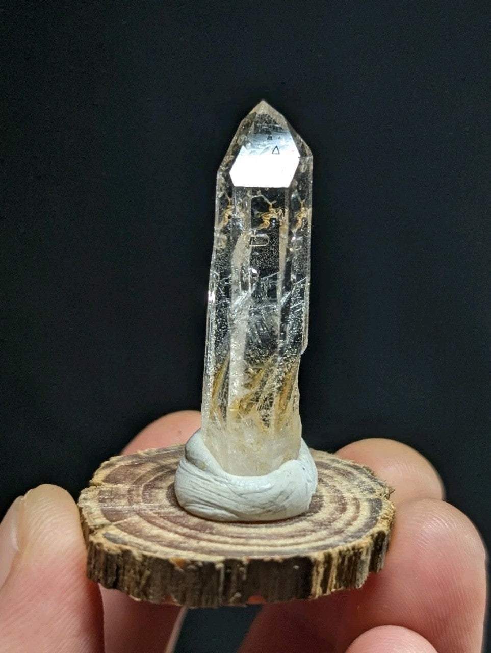 A clear, translucent quartz crystal with a pointed tip and visible internal inclusions sits atop a circular wooden slice base.