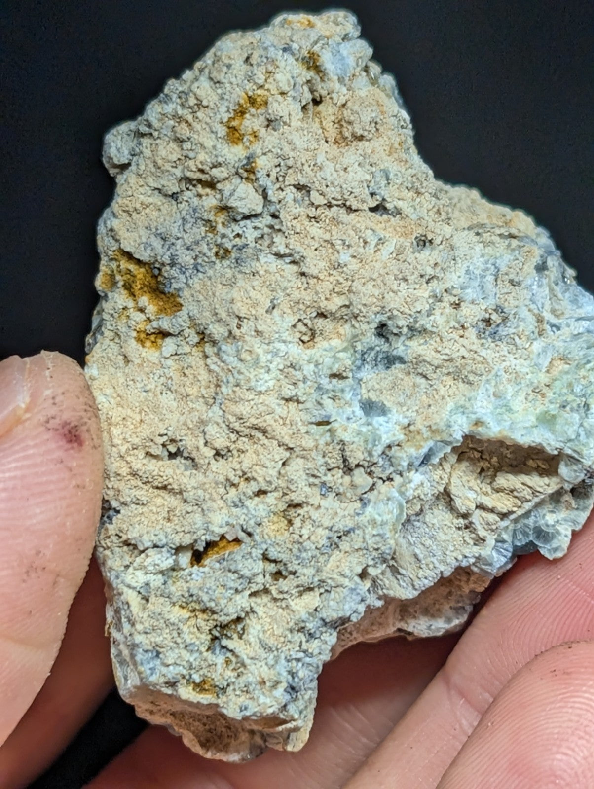 A rough, irregularly shaped mineral specimen with a mottled surface of pale blue-green and beige tones, accented by patches of yellowish discoloration.