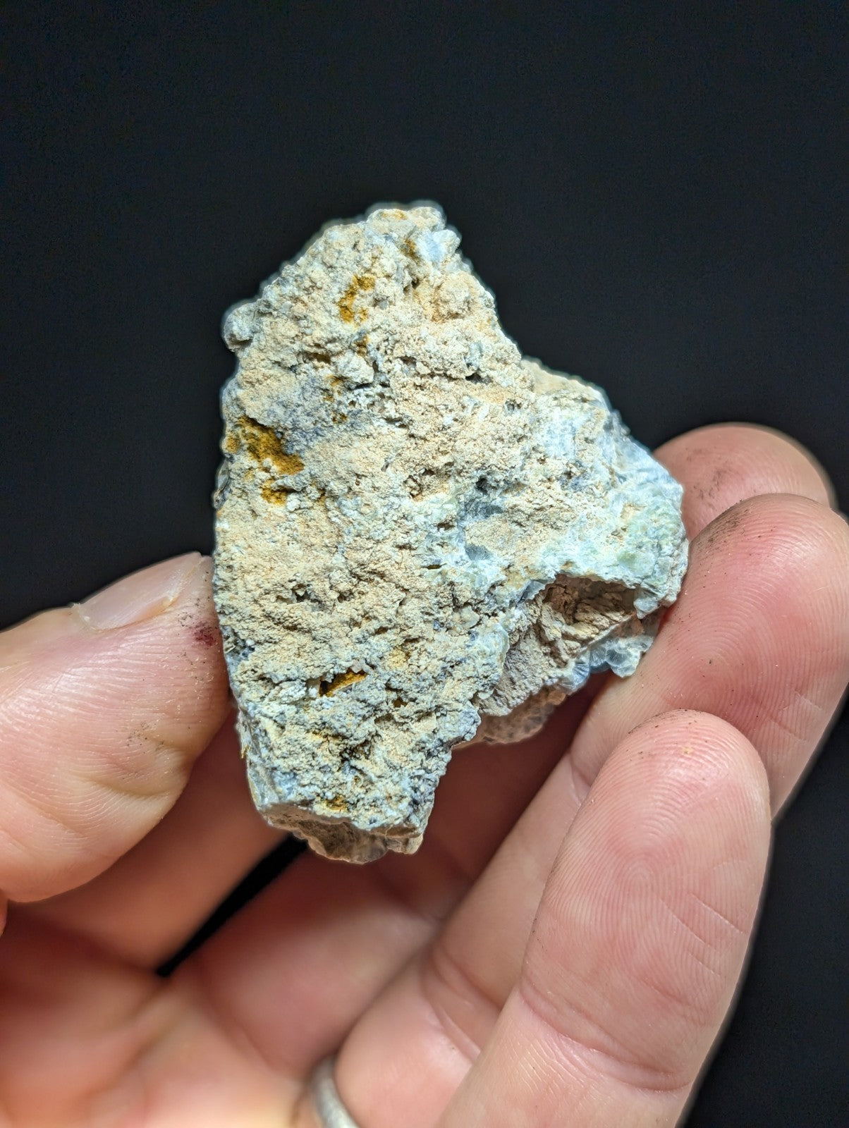 A rough, irregularly shaped mineral specimen with a mottled surface of light blue-green and beige tones, accented by patches of yellowish-orange.