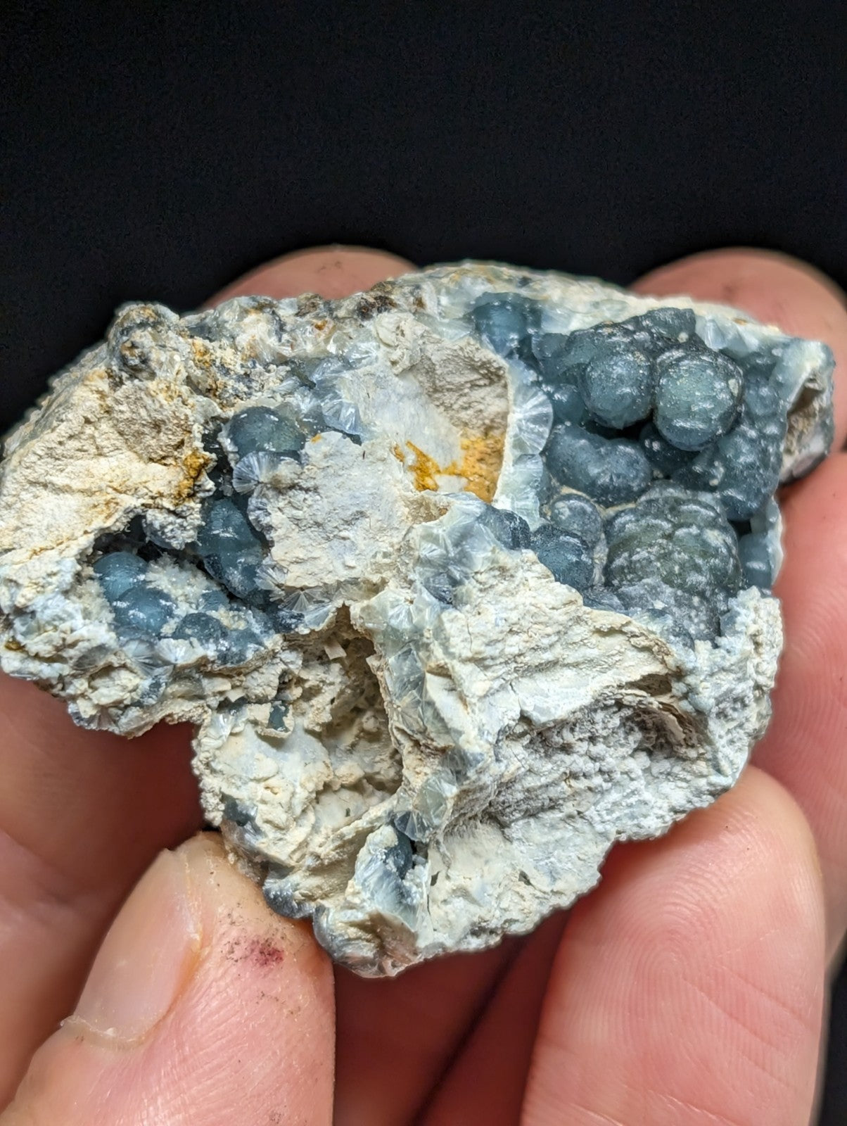 A hand holds a natural mineral specimen featuring clusters of translucent blue-gray spherical crystals embedded in a rough, light gray matrix with subtle yellowish patches.