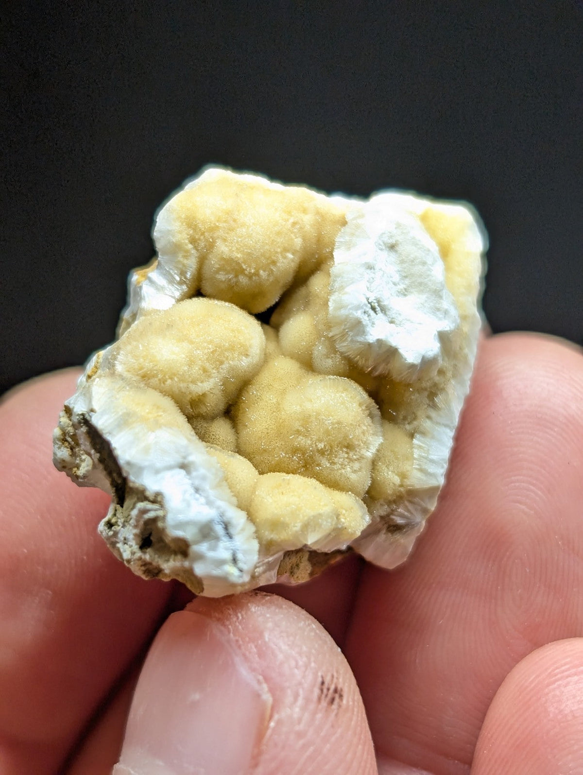A hand holds a small, irregularly shaped mineral specimen featuring clusters of pale yellow and white crystalline formations with rough, textured surfaces.