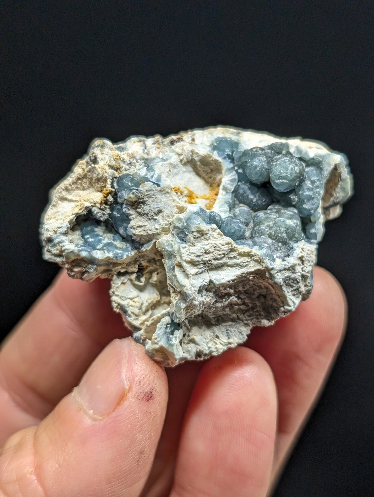 A hand holds a rough, irregularly shaped mineral specimen featuring clusters of translucent blue crystals embedded in a pale gray matrix.