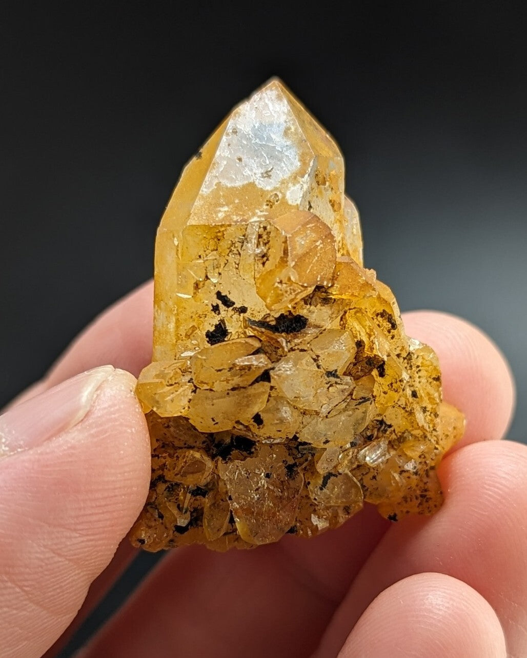 A translucent golden-yellow quartz crystal cluster with a pointed tip and embedded dark inclusions, held gently between fingers.