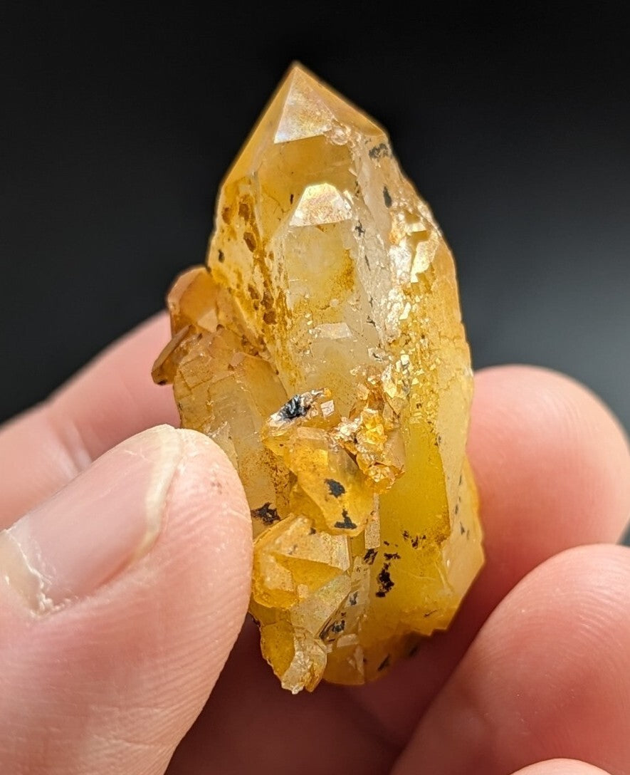 A translucent golden-yellow crystal with a pointed tip and embedded dark inclusions, held gently between fingers.
