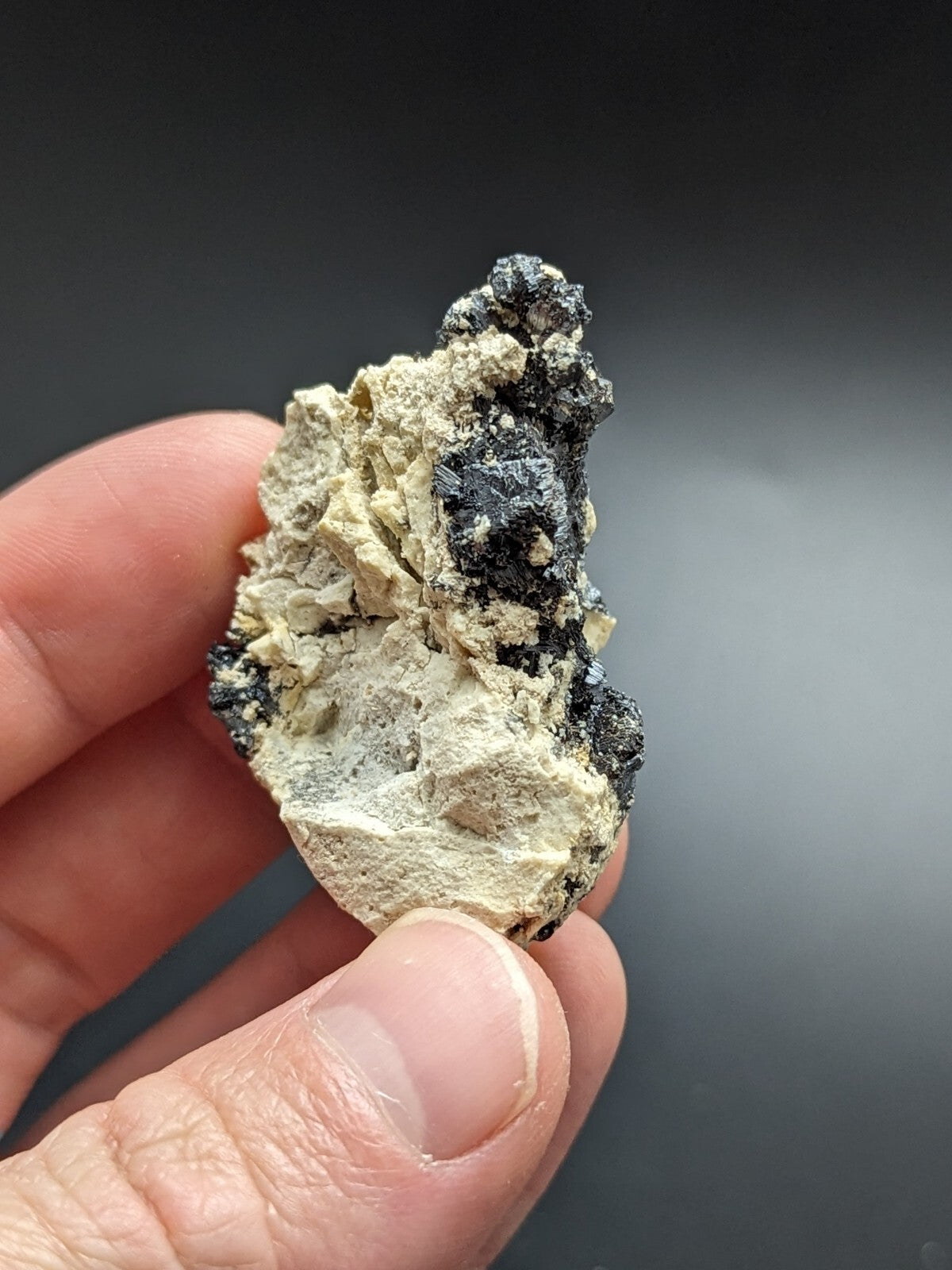 A hand holds a rough, irregularly shaped mineral specimen featuring patches of dark black and light beige material.