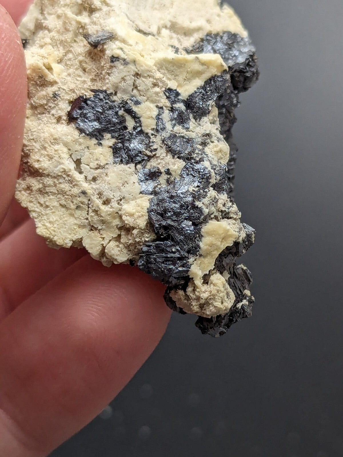 A hand holds a rough, irregularly shaped mineral specimen featuring patches of dark metallic luster contrasting with its pale yellowish matrix.