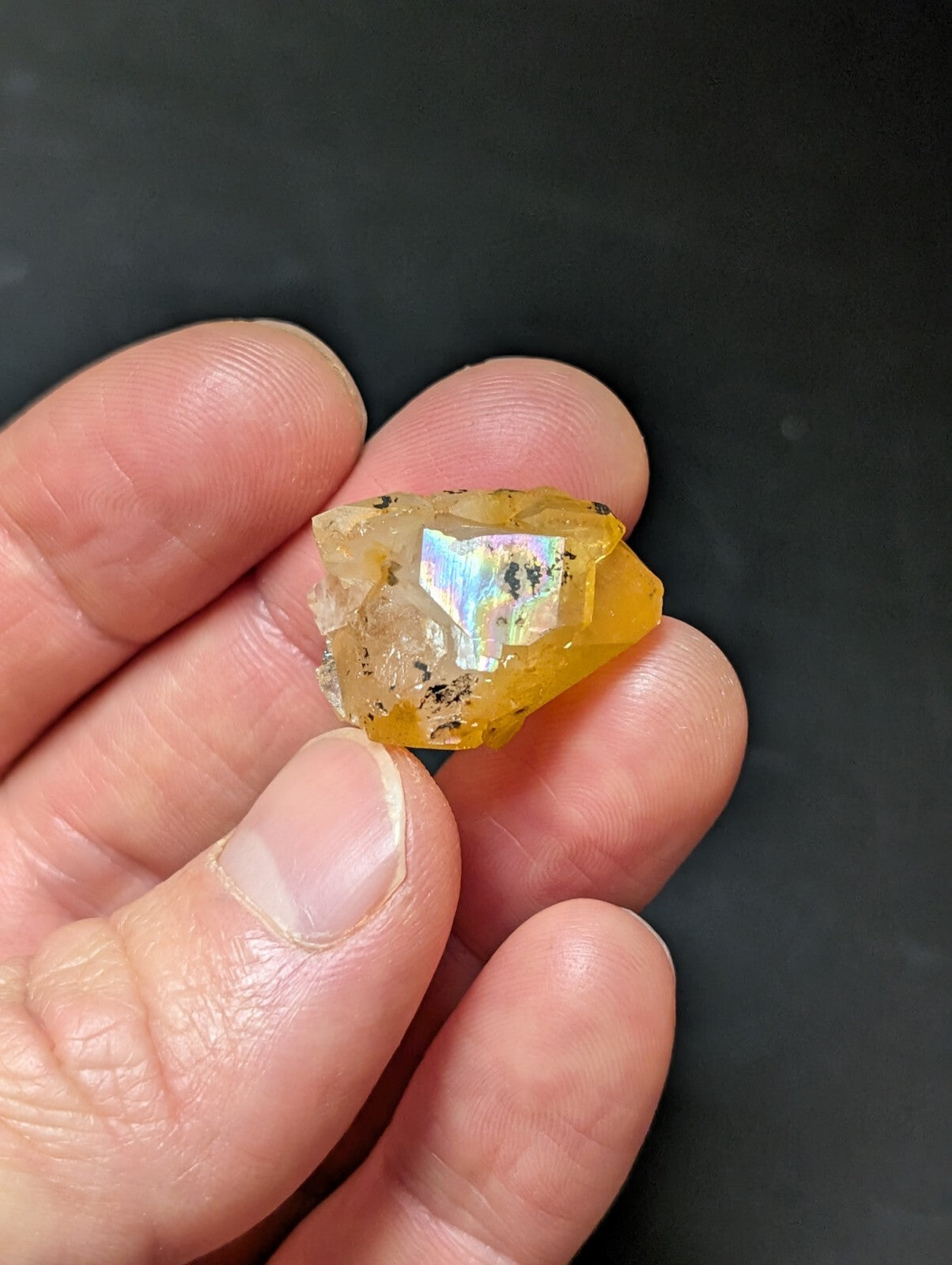 A translucent yellow crystal with iridescent facets and dark inclusions, held gently between fingers.