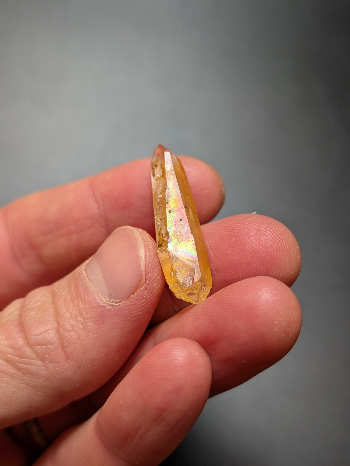 A translucent golden-yellow crystal with a pointed tip and subtle iridescent flashes, held delicately between fingers.