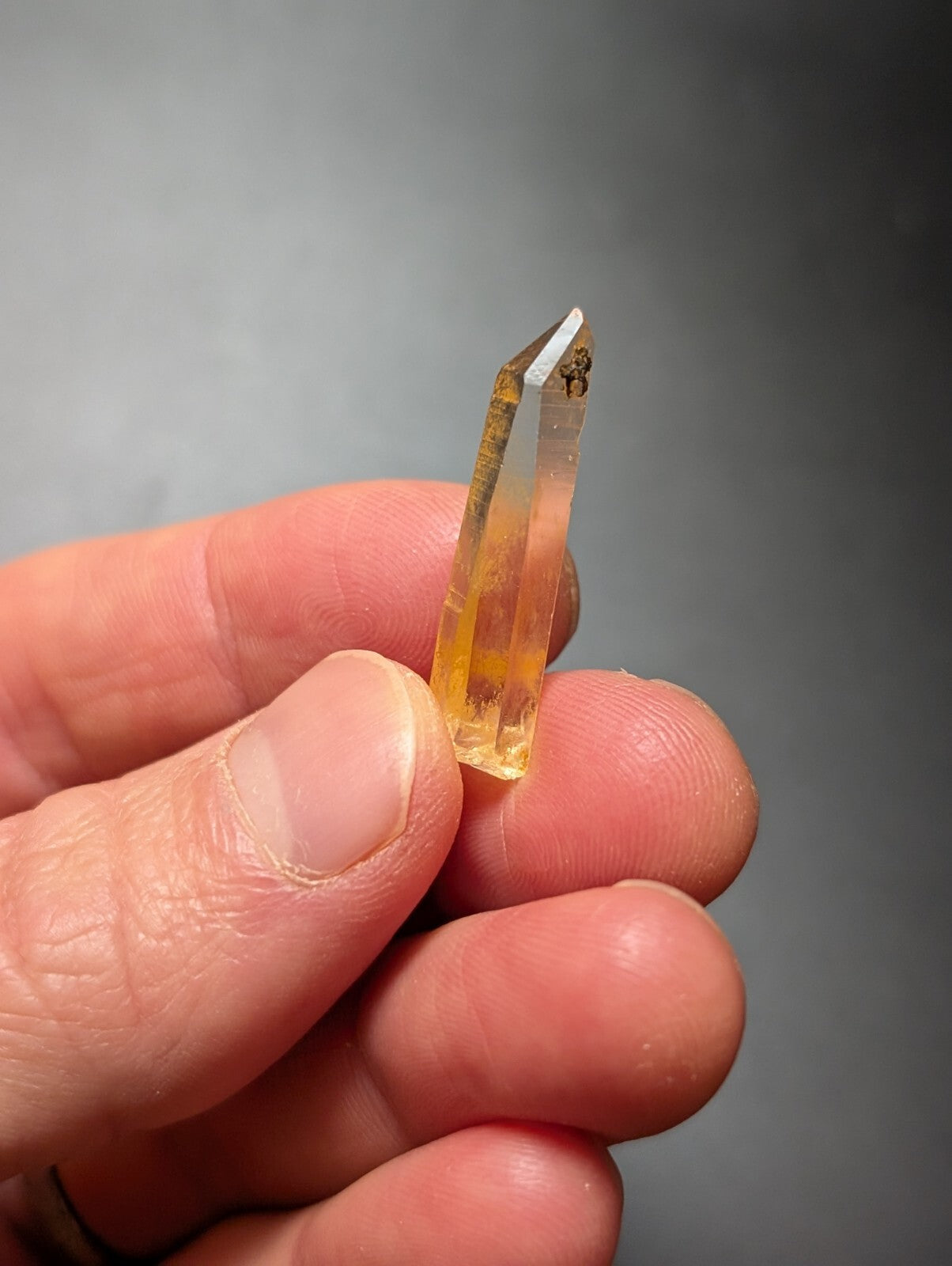 A translucent yellow-orange crystal with a pointed tip, held delicately between fingers.