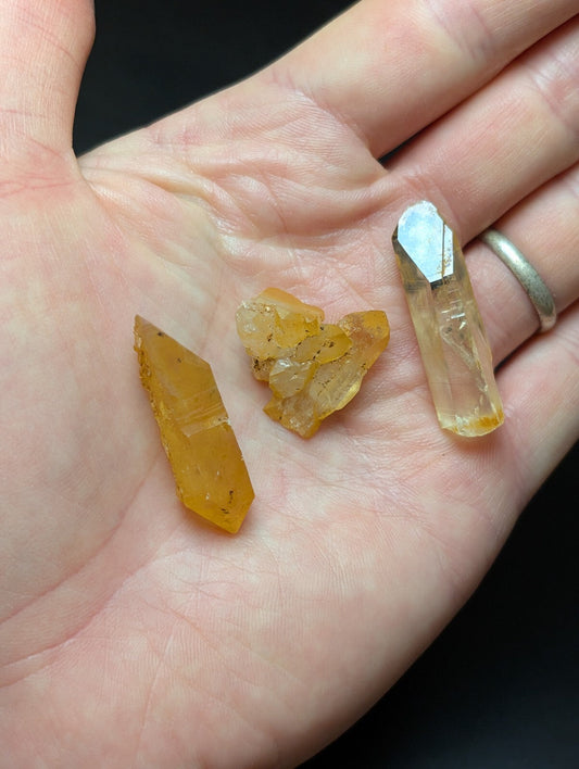 A hand holds three distinct yellow-orange crystal specimens, including a faceted point and an irregular cluster.