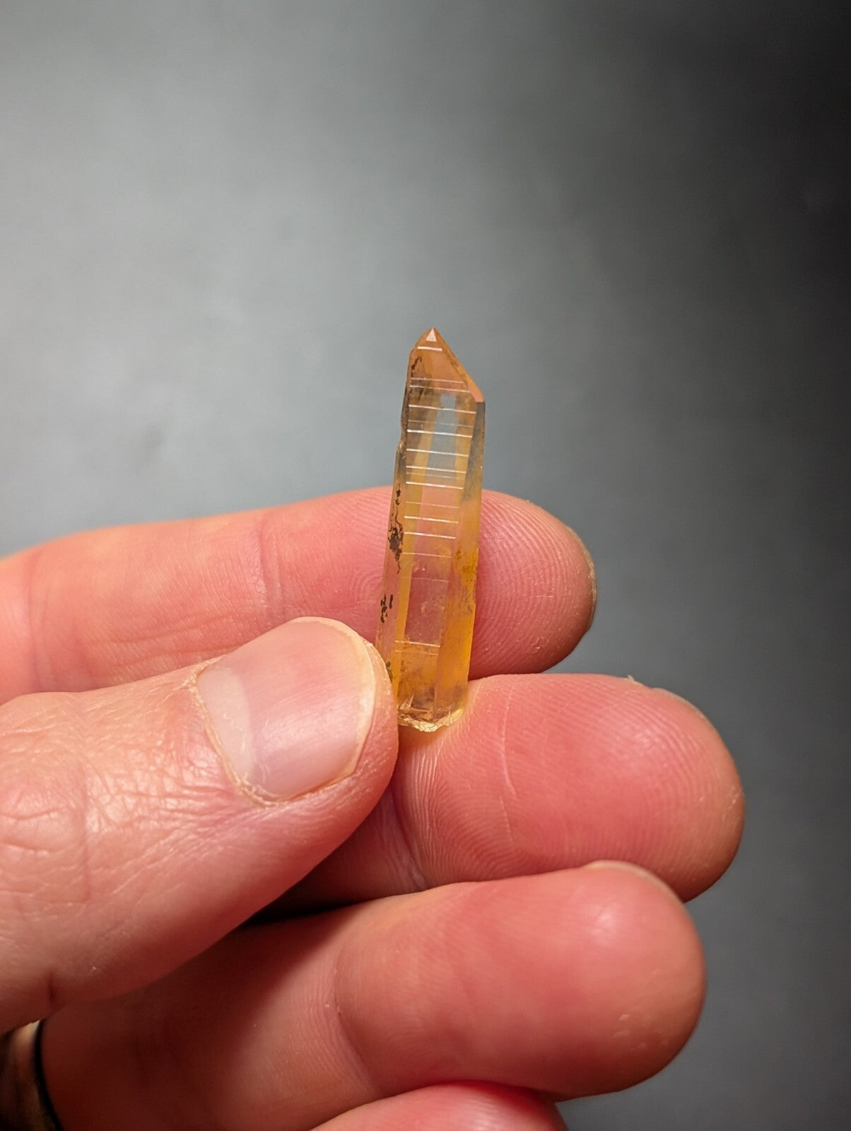 A translucent amber-colored quartz crystal with a pointed tip and visible internal striations, held delicately between fingers.