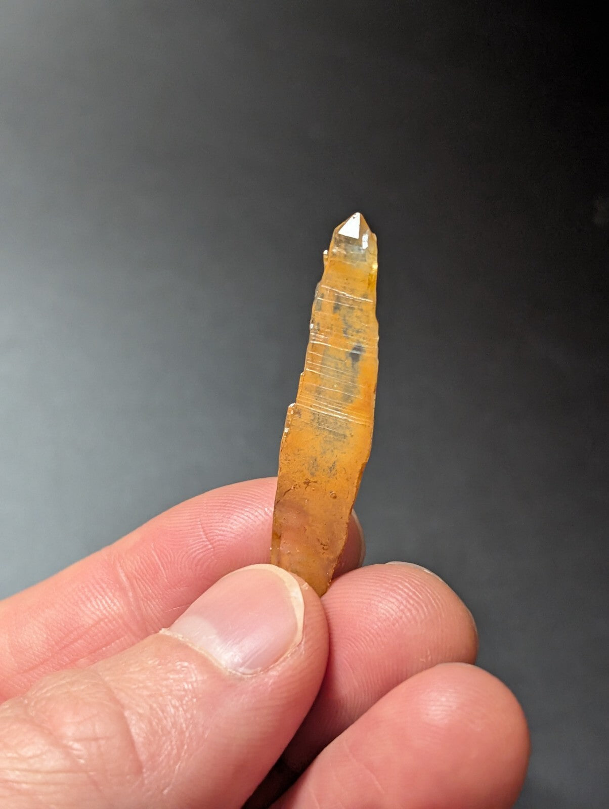A translucent amber-colored quartz crystal with a pointed tip and visible striations, held gently between fingers.