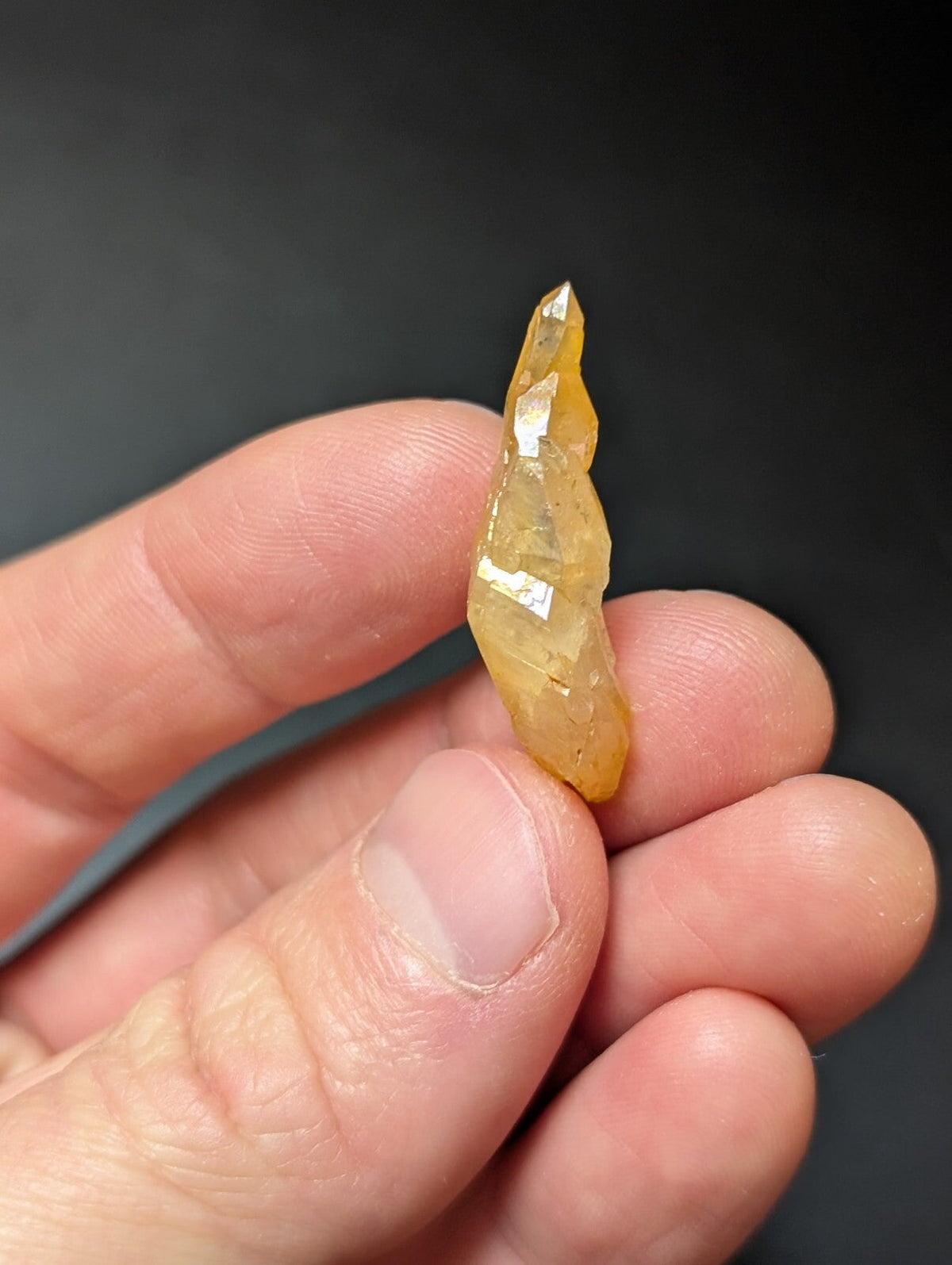 A translucent yellow crystal with a pointed tip and faceted surface, held delicately between fingers.