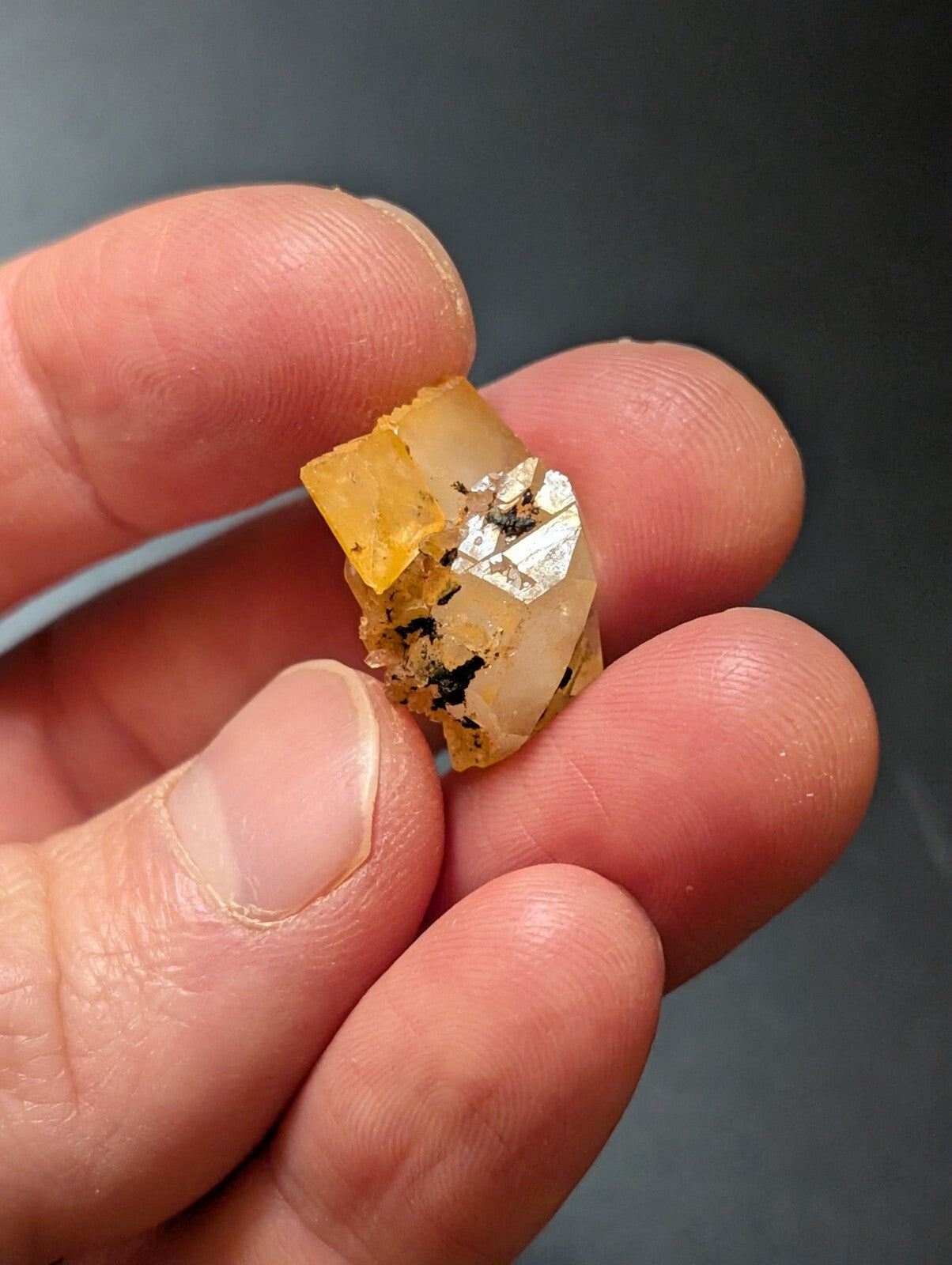 A translucent yellowish mineral specimen with a metallic silver inclusion and dark speckles, held delicately in someone’s hand.