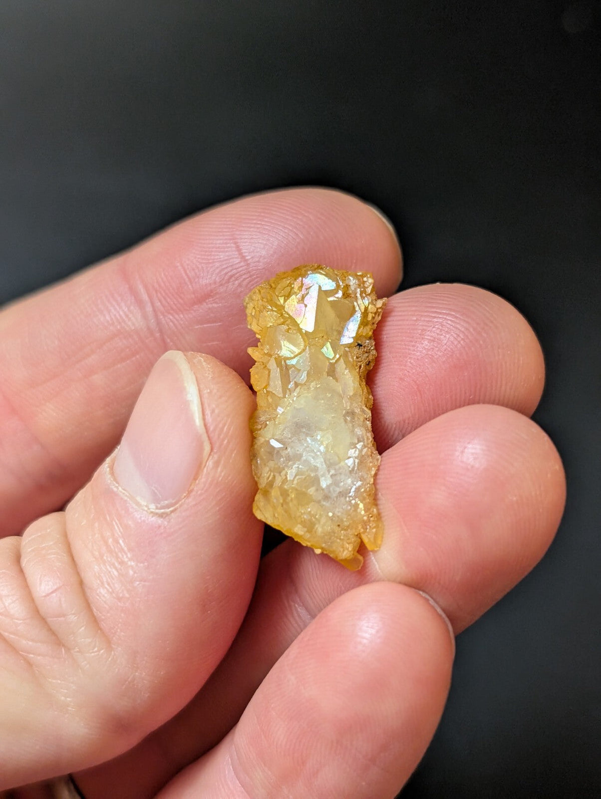 A translucent golden-yellow crystal with a rough, jagged surface and iridescent facets held in someone’s hand.
