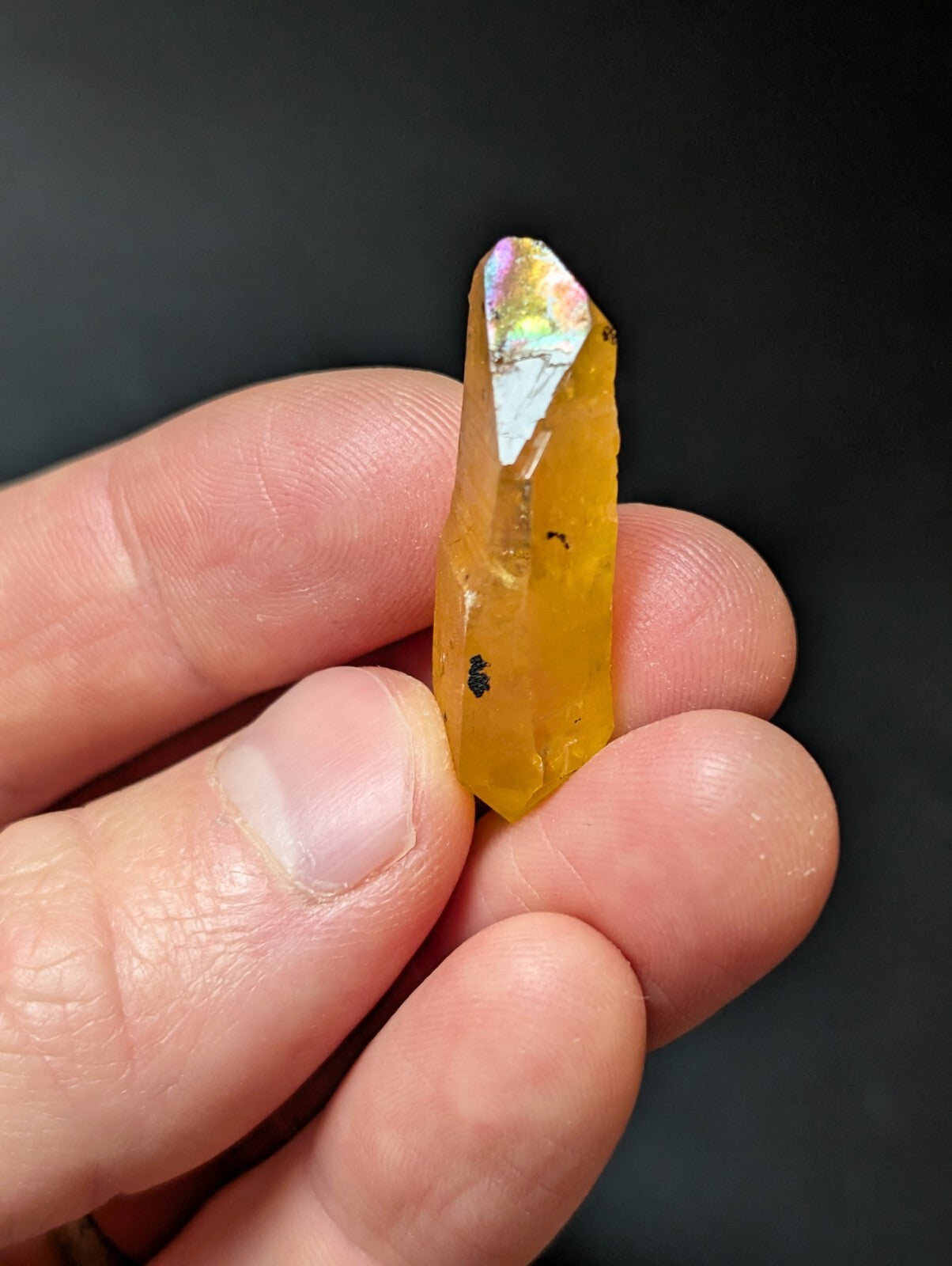 A translucent golden-yellow crystal with a pointed, faceted tip and iridescent rainbow hues at its top.