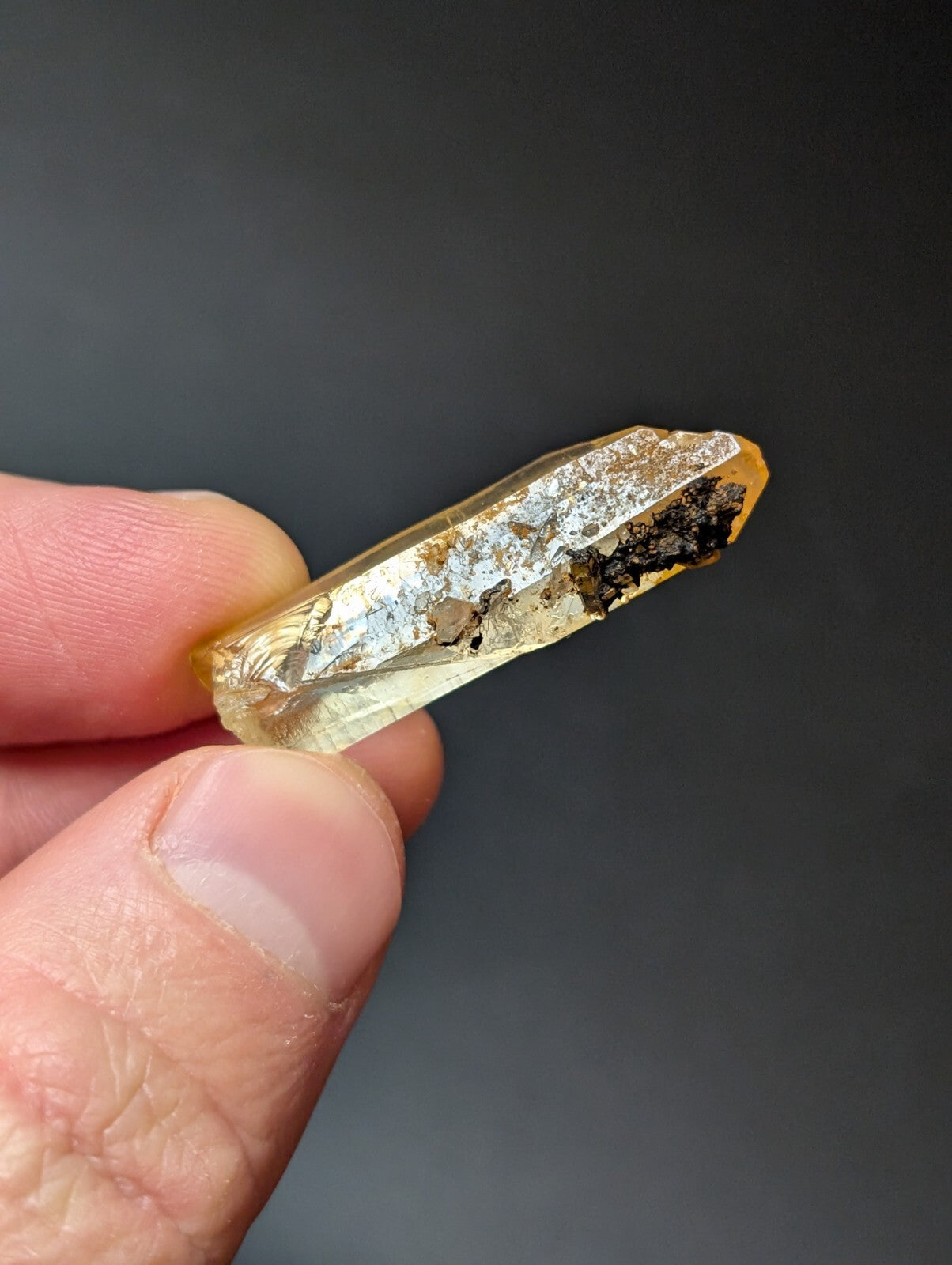 A translucent golden-yellow quartz crystal with a pointed tip and natural inclusions, held between fingers against a dark background.