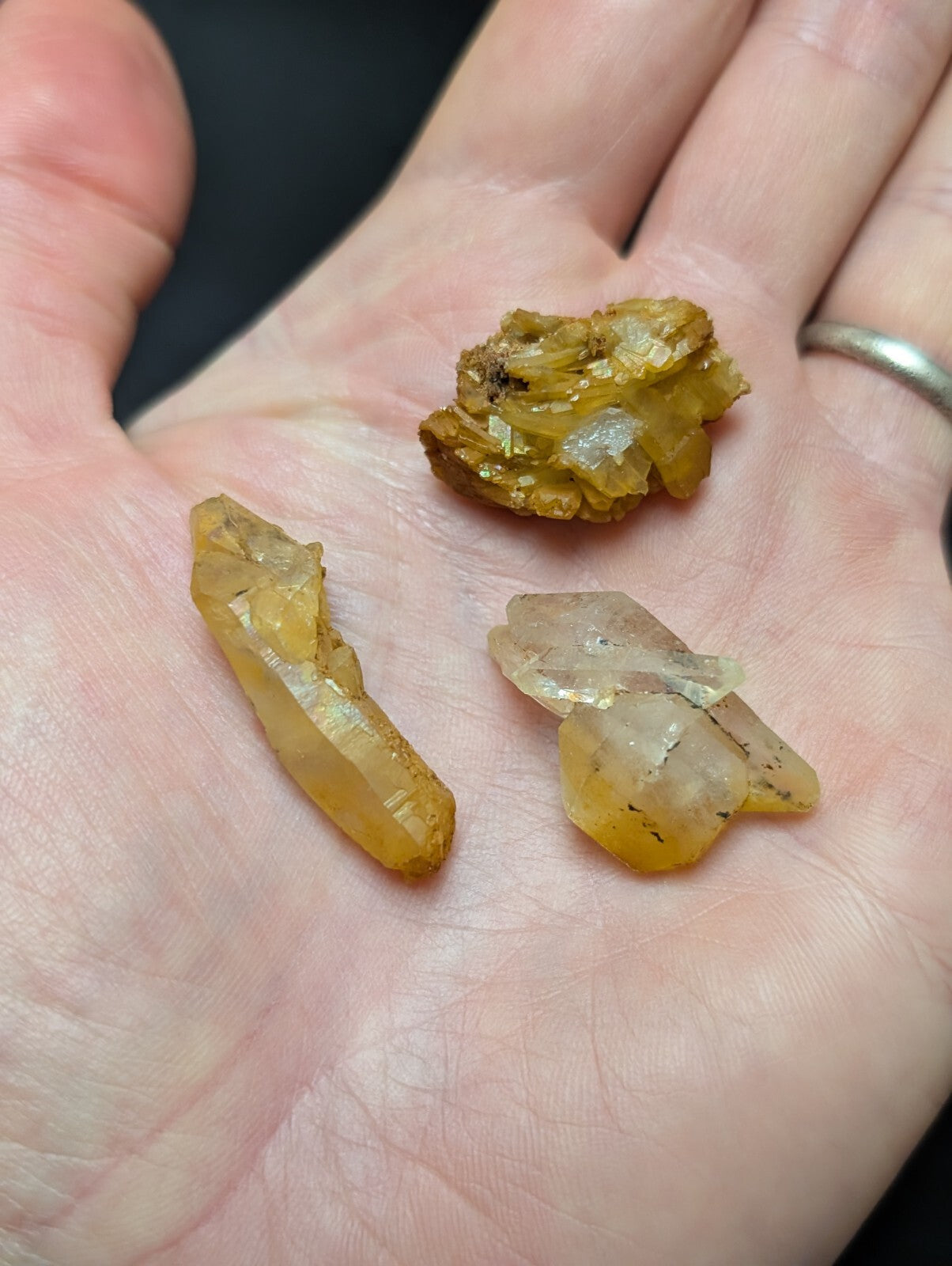 A hand holds three distinct yellowish-orange mineral crystals, featuring translucent and semi-translucent facets with natural imperfections.