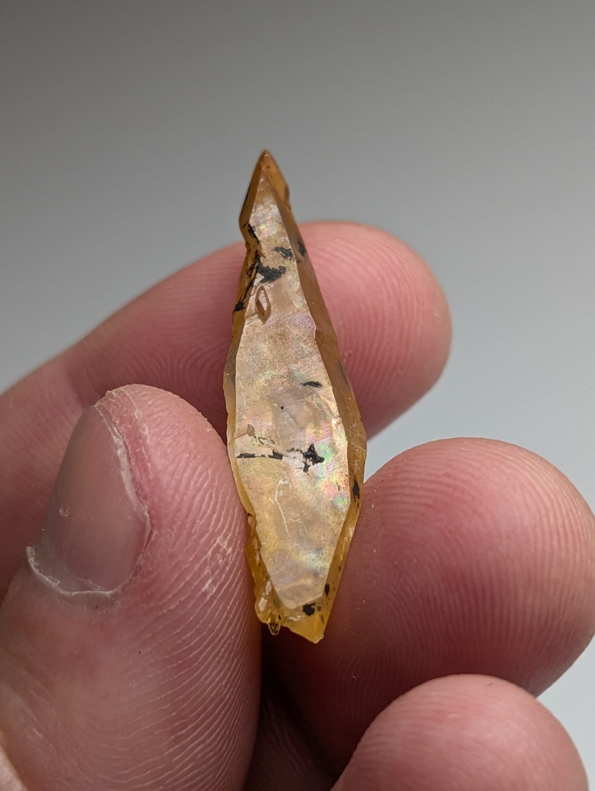 A translucent amber arkansas quartz crystal with natural iridescence and black inclusions held between fingers
