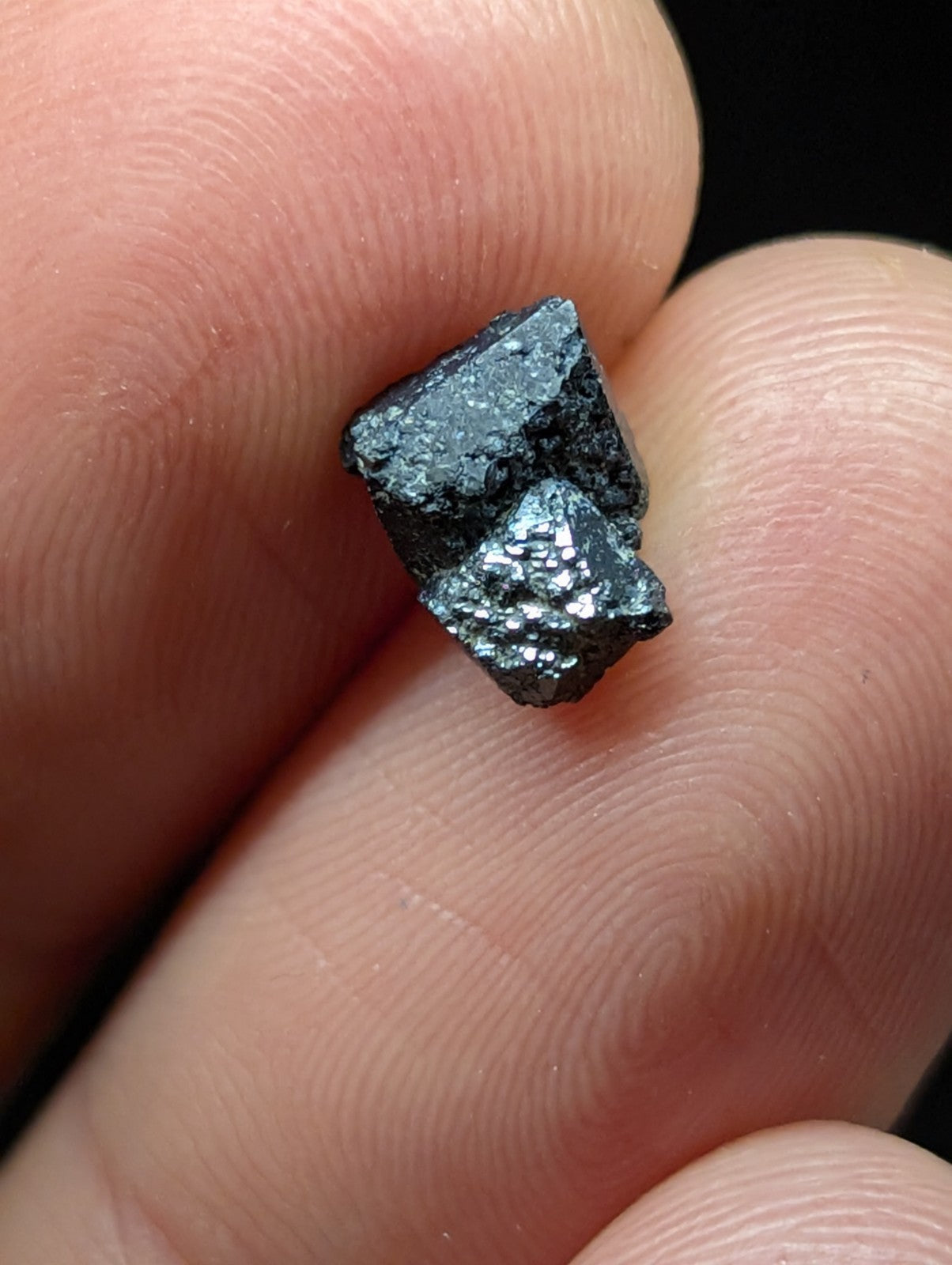 A small, dark metallic mineral specimen with a rough, faceted surface and subtle iridescent sheen.