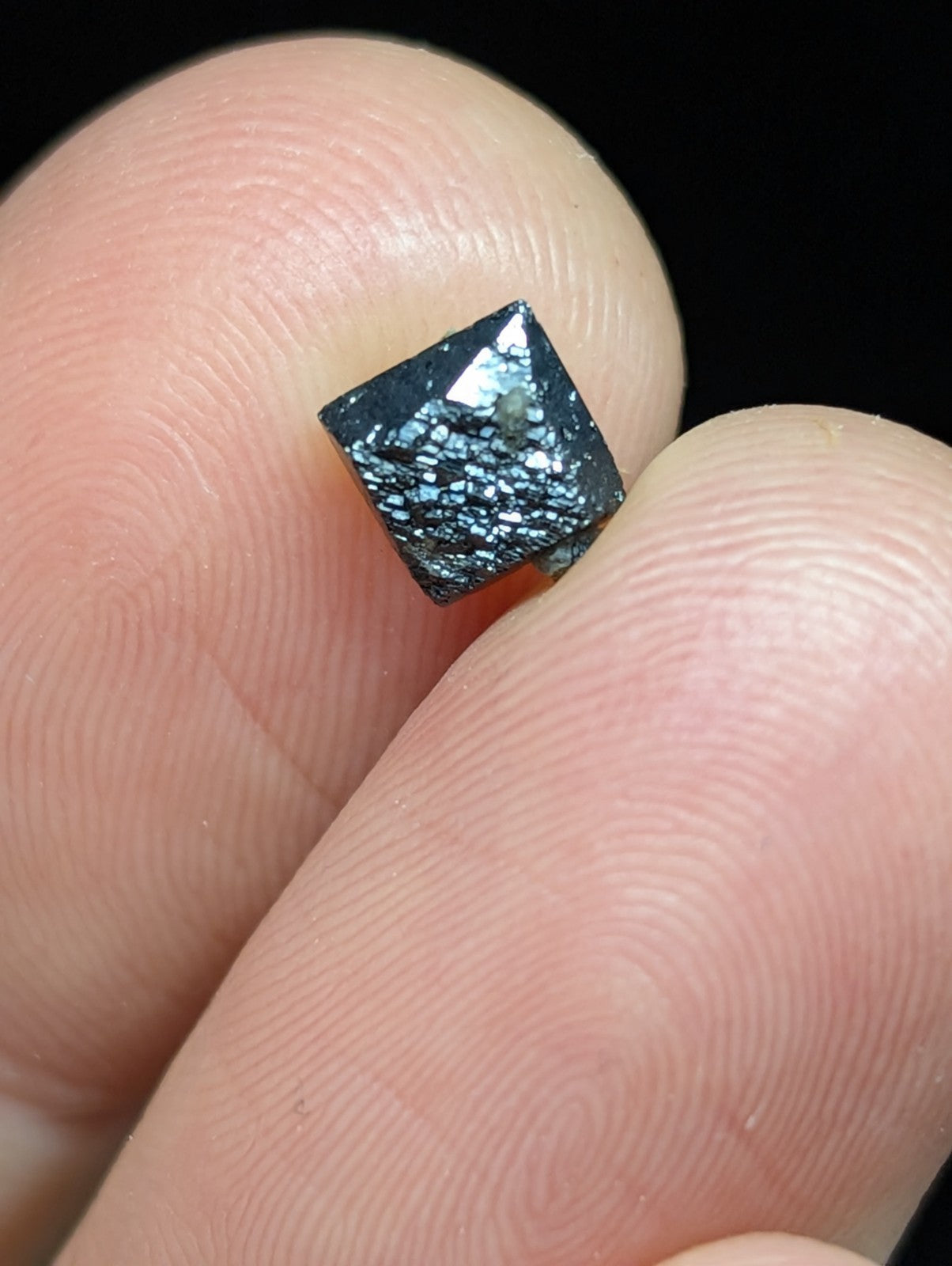 A dark, square-cut gemstone with a shimmering blue and black speckled surface held between fingers.
