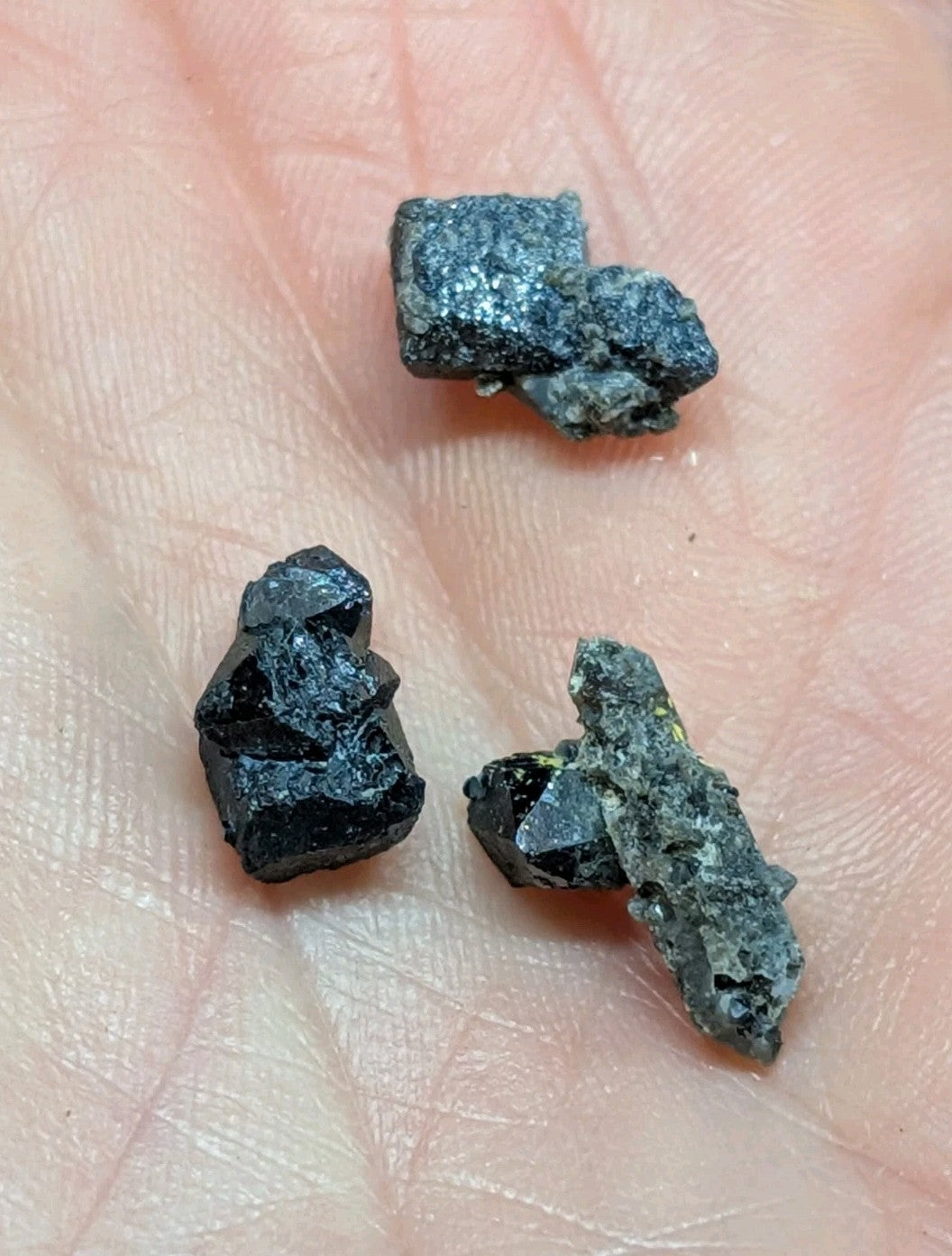 Three rough, dark mineral specimens with jagged edges and a matte black to deep gray surface texture rest in the palm of a hand.