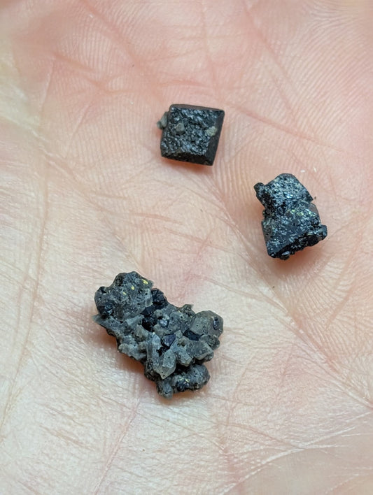 Three irregularly shaped, dark gray to black mineral fragments with rough, textured surfaces rest in the palm of a hand.