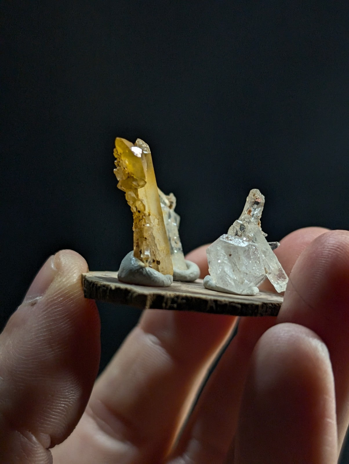 A hand holds a miniature crystal display featuring two distinct mineral specimens—one with translucent amber-yellow hues and the other clear, both mounted on a rustic wooden base.