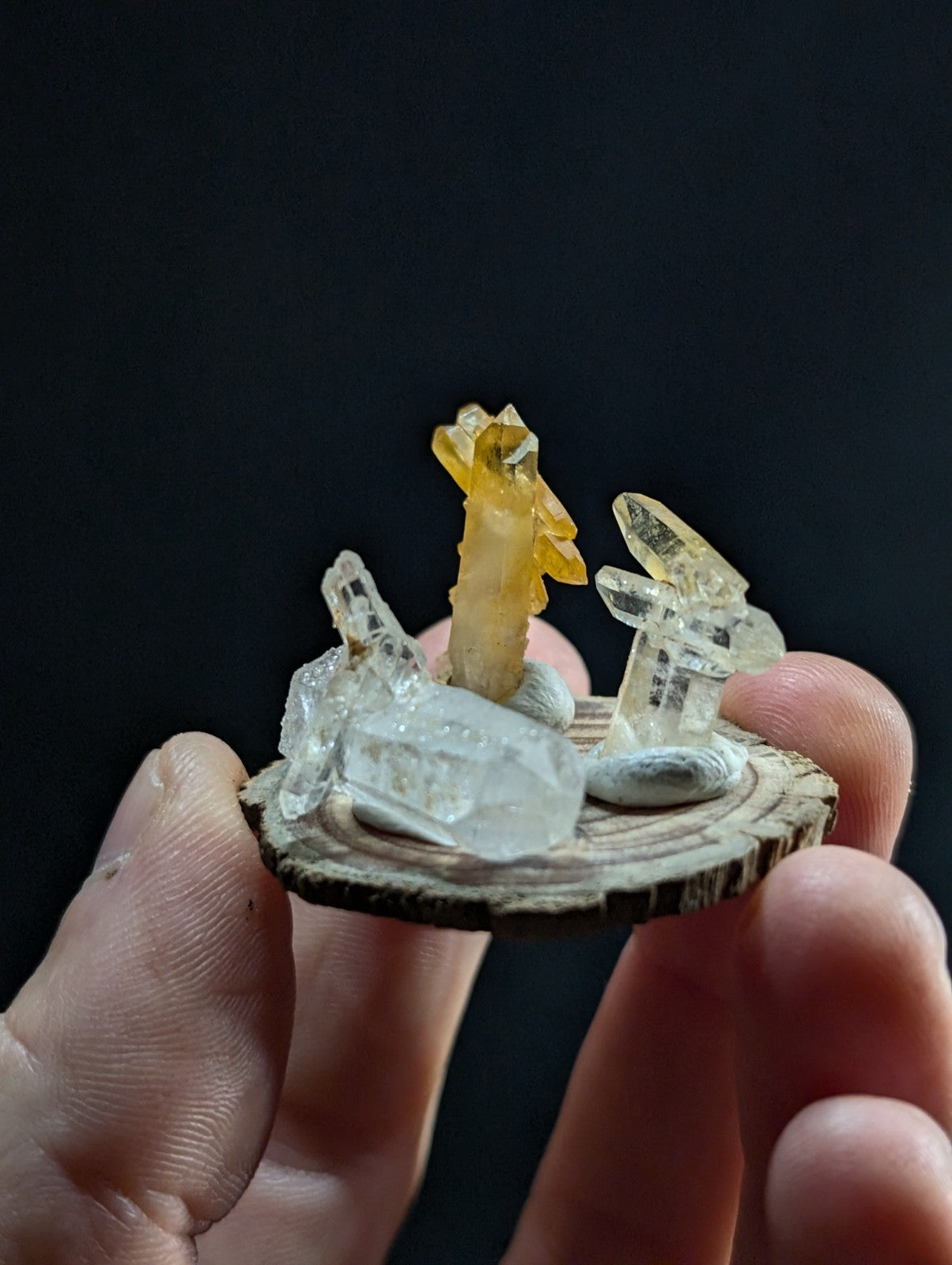 A miniature crystal sculpture featuring a golden-yellow citrine figure and clear quartz formations mounted on a rustic wooden slice base.