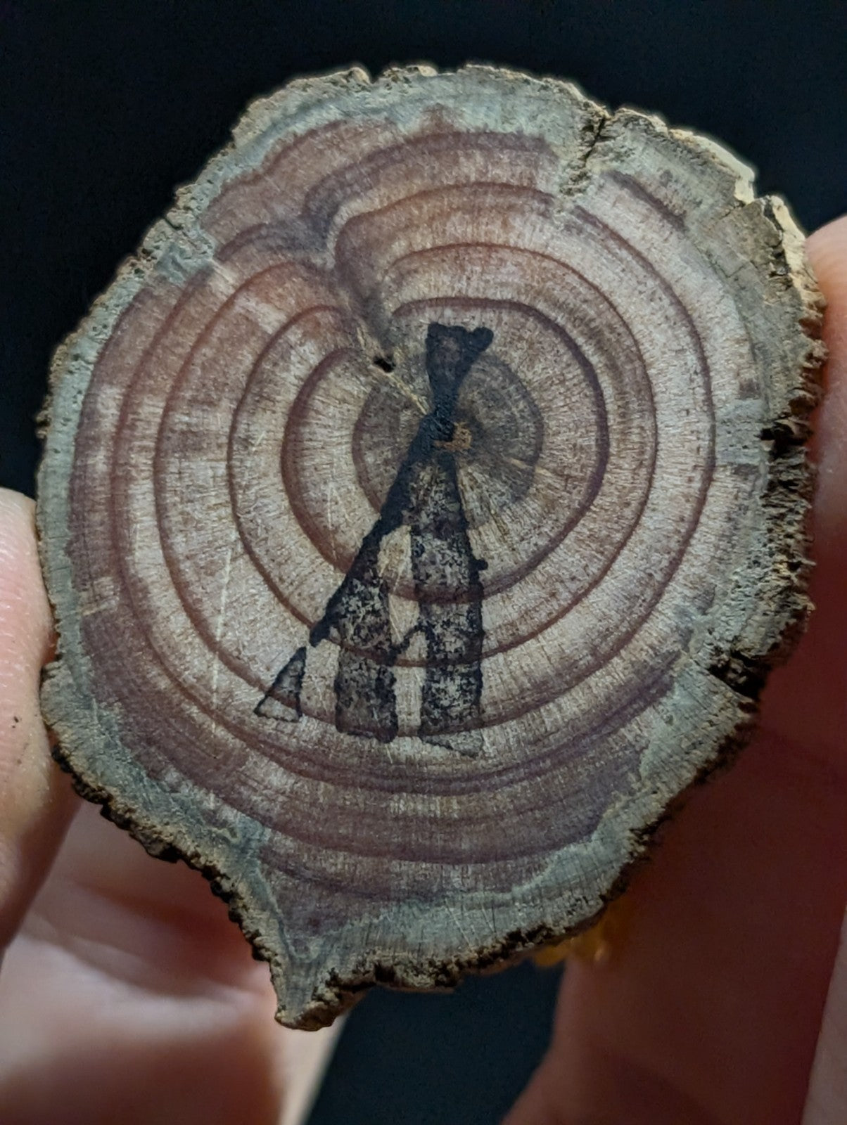 A circular wooden slice with natural concentric rings and a dark, hand-painted silhouette of a figure in the center.