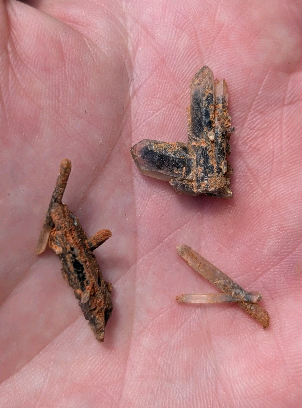 A hand holds three distinct mineral specimens: a rusted, angular iron fragment with visible corrosion; a larger quartz crystal cluster exhibiting clear transparency and dark inclusions; and two slender, translucent quartz needles.