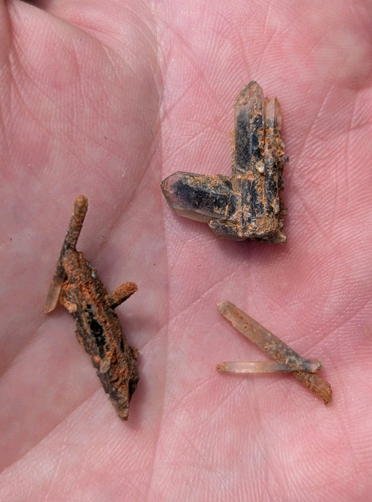 A hand holds three distinct mineral specimens: a rusted, angular iron fragment with visible corrosion; a larger quartz crystal cluster exhibiting clear transparency and dark inclusions; and two slender, translucent quartz needles.