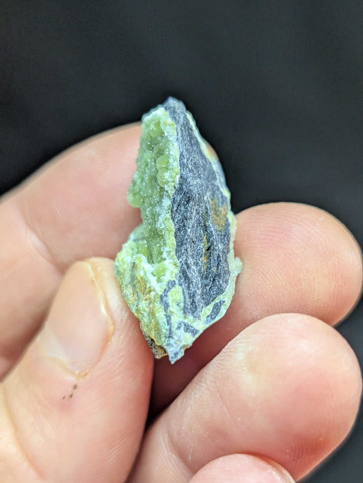 A hand holds a small, irregularly shaped mineral specimen featuring a vibrant green crystalline formation alongside darker grayish-black areas.