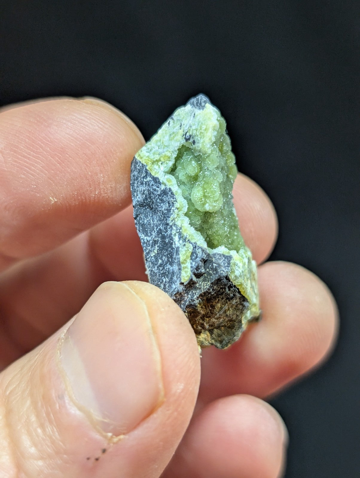 A hand holds a natural green mineral specimen with crystalline formations and dark gray matrix.