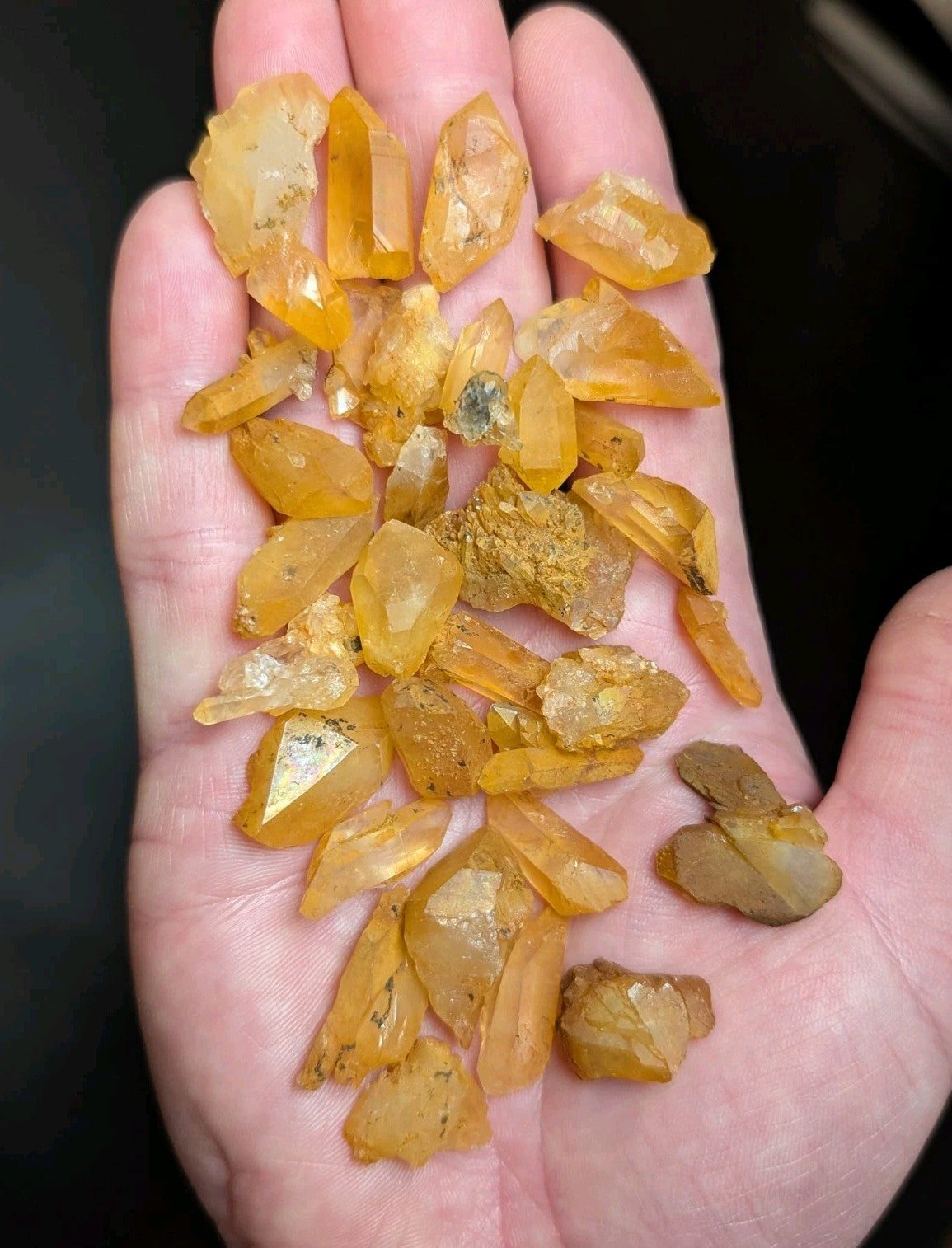 A hand holds a collection of translucent yellow-orange crystal fragments with jagged, faceted edges and some embedded dark inclusions.