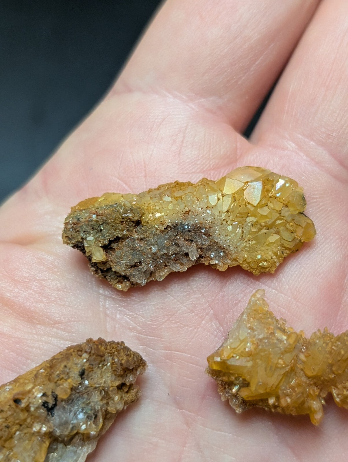 A hand holds three irregularly shaped, golden-yellow mineral specimens with translucent crystalline formations and earthy brown matrix.